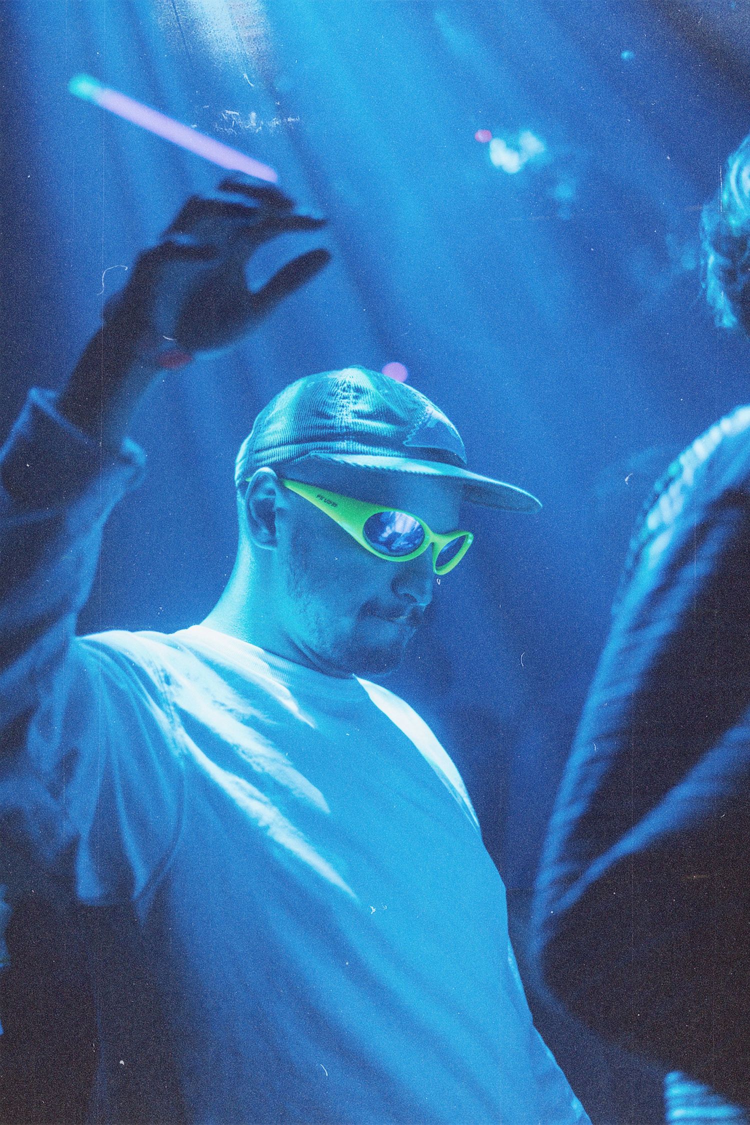 Man wearing green sunglasses and a cap under blue neon lights at a nightclub event.