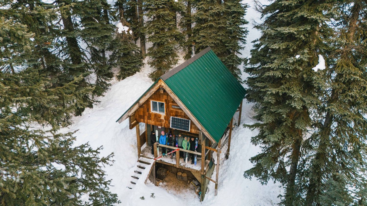 Aerial view of a cozy wooden cabin with green roof surrounded by snow-covered pine trees.