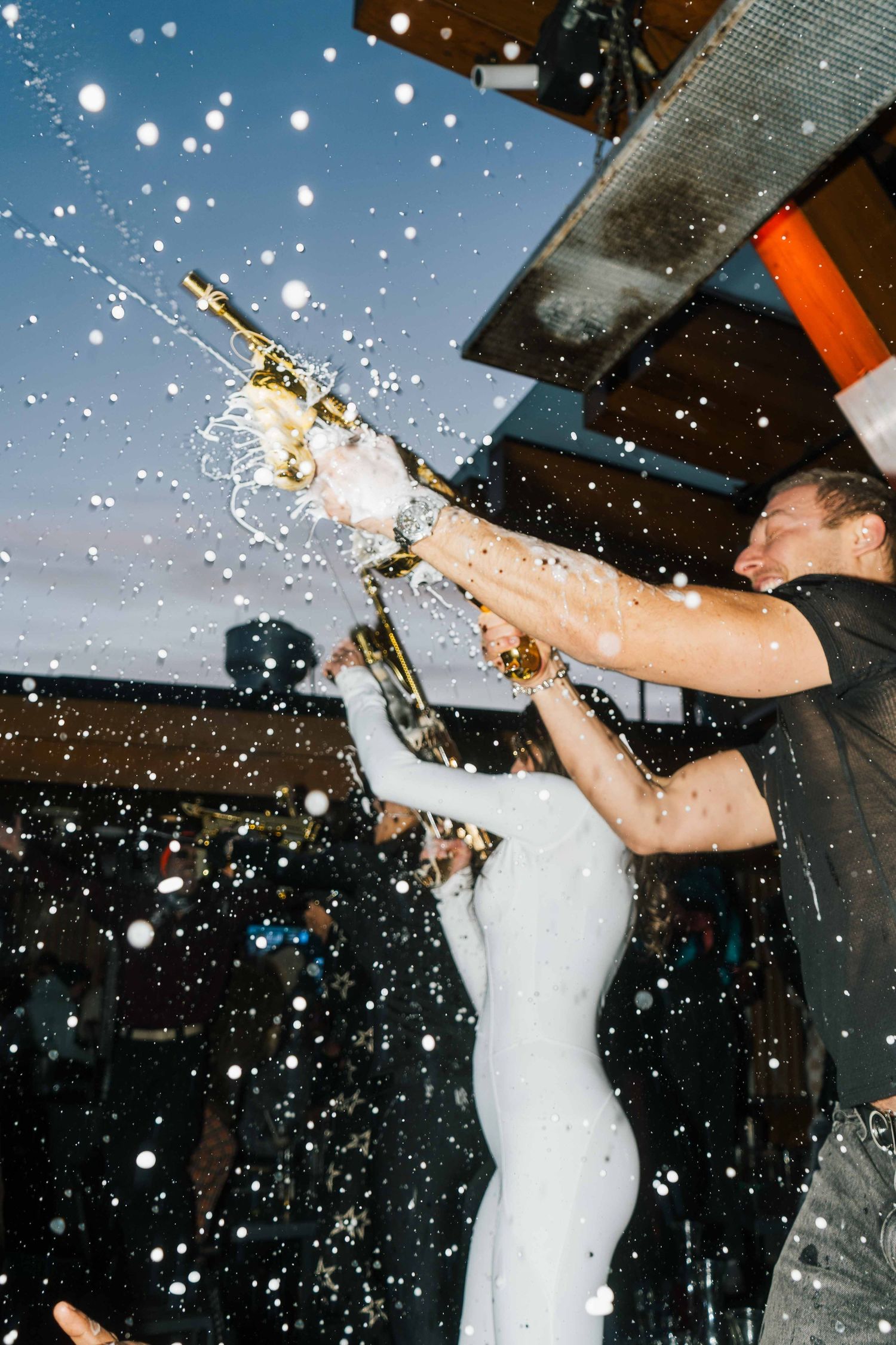 Bride and groom spraying champagne bottles in celebration at an outdoor evening wedding reception.