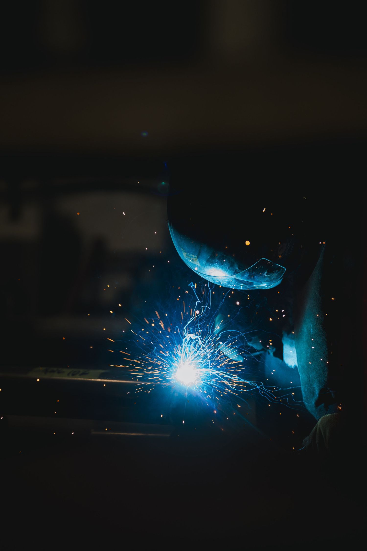 Welder working in a dark workshop with bright blue sparks flying from an arc welding torch.