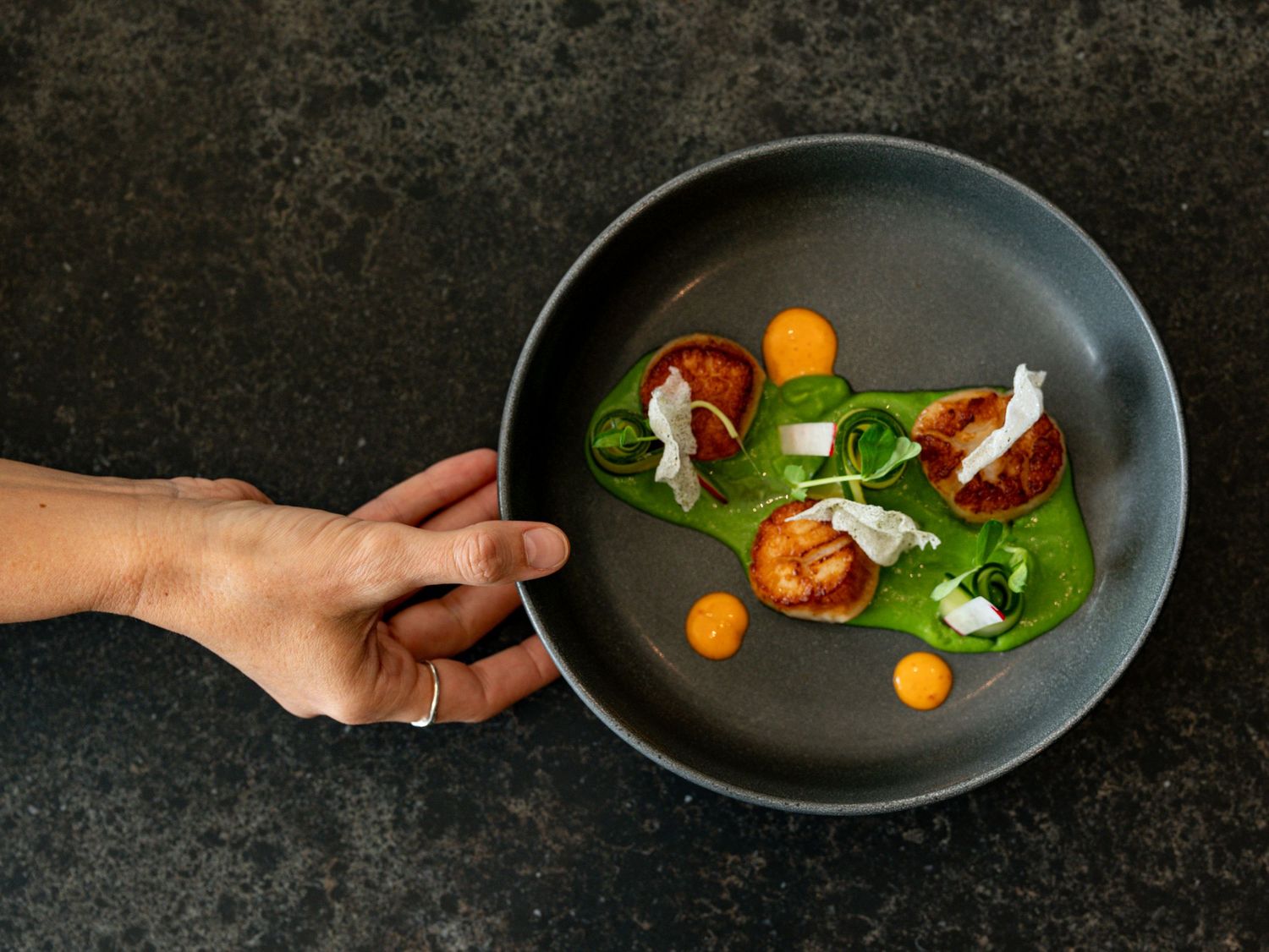 Hand holding a dark plate with artistic fine dining dish featuring tomatoes and greens.