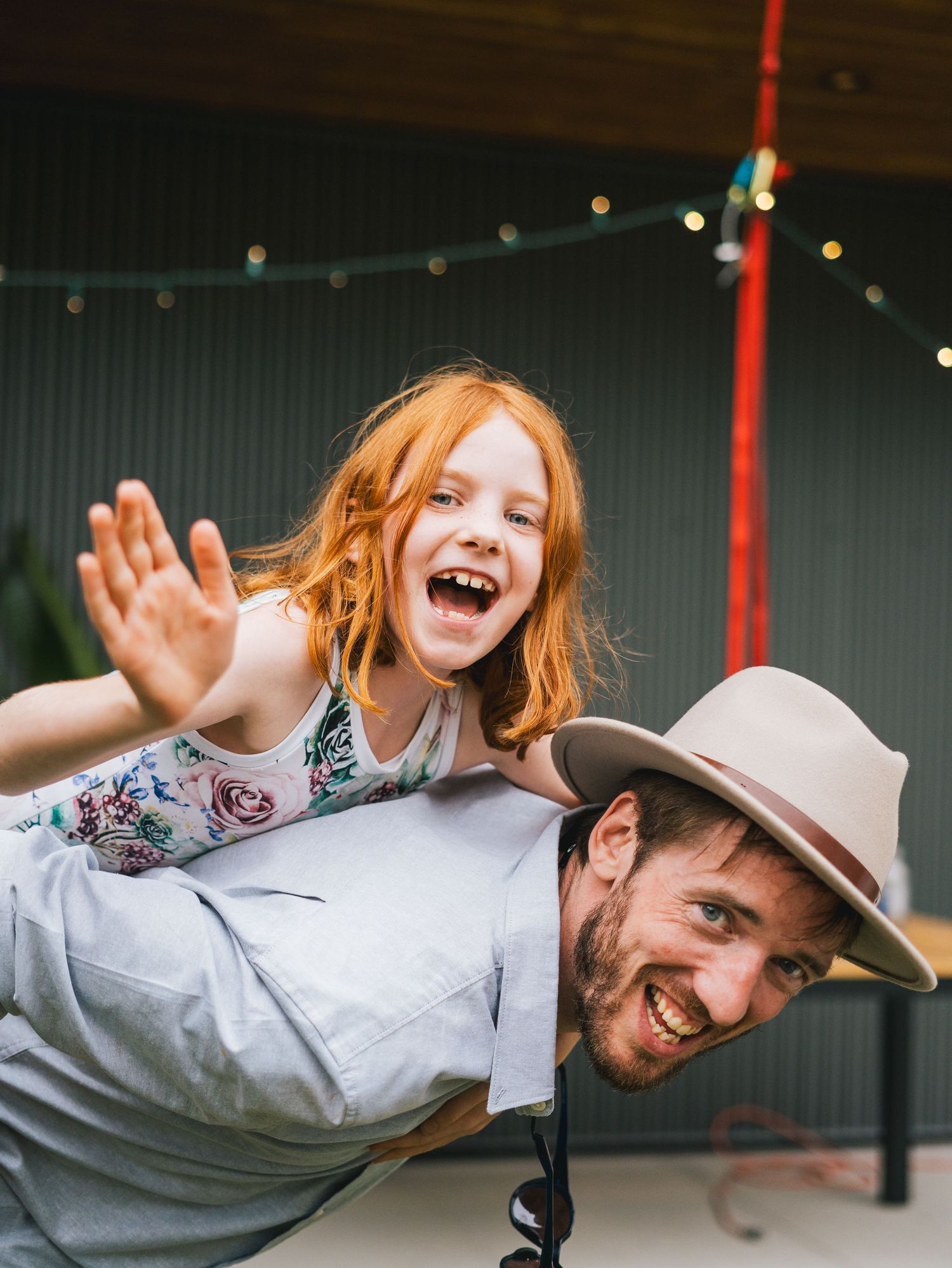 Laughing man in a hat gives a joyful red-haired girl a piggyback ride under string lights outdoors.
