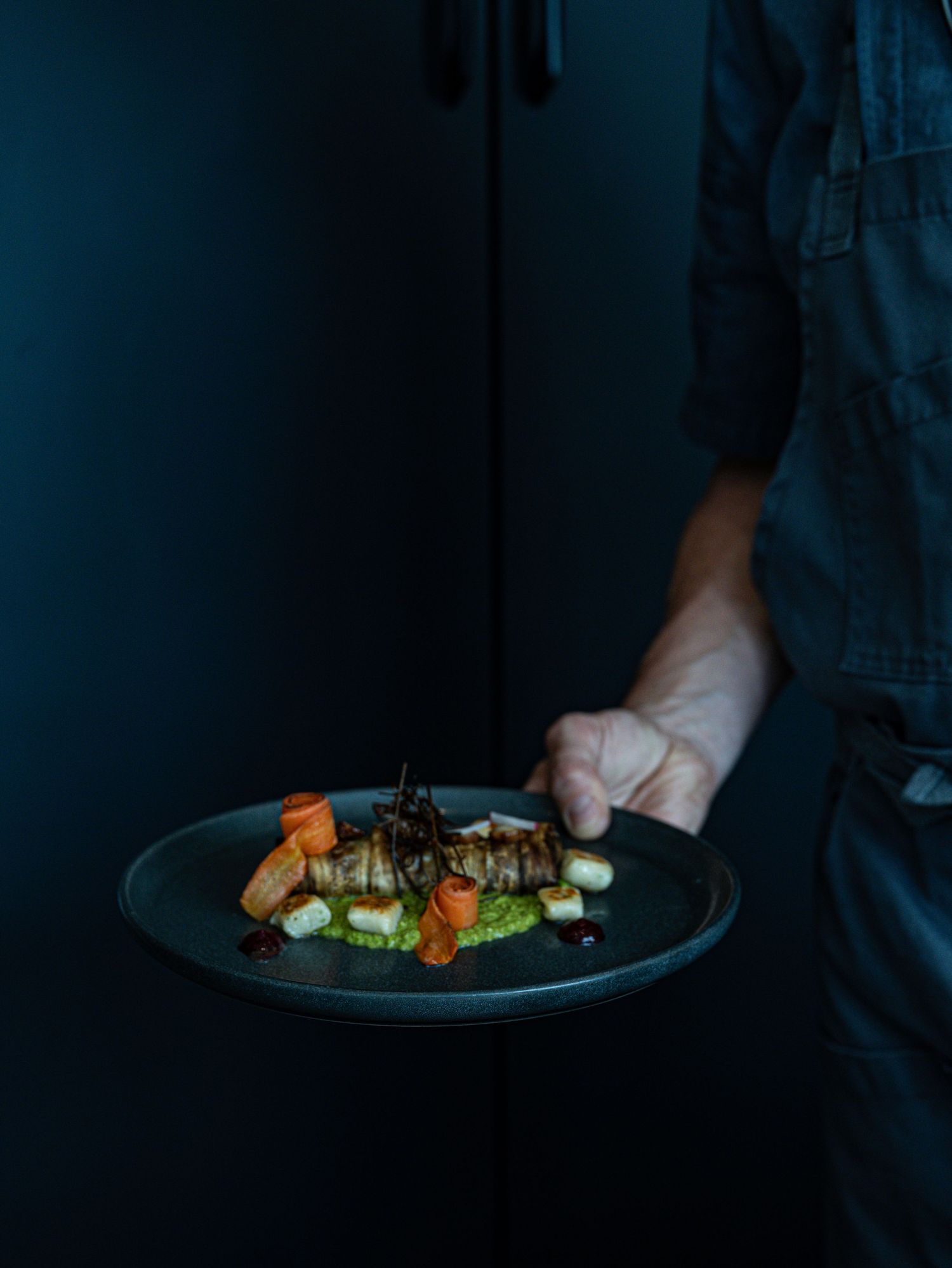 Chef holding a dark plate with an elegantly plated gourmet dish featuring vegetables and sauce.