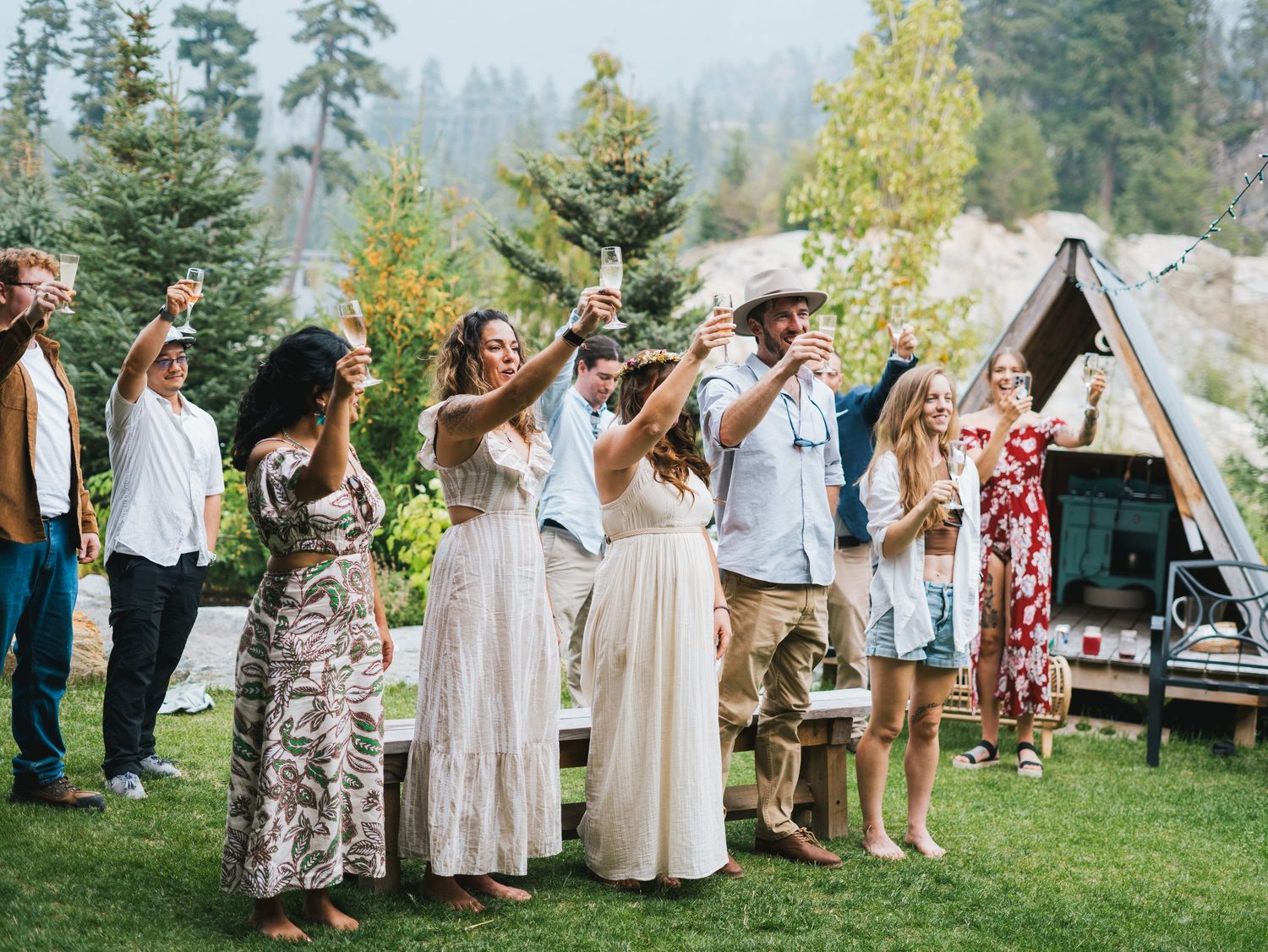 Group of friends raising glasses in a toast outdoors near a forest with a glamping tent in the background.