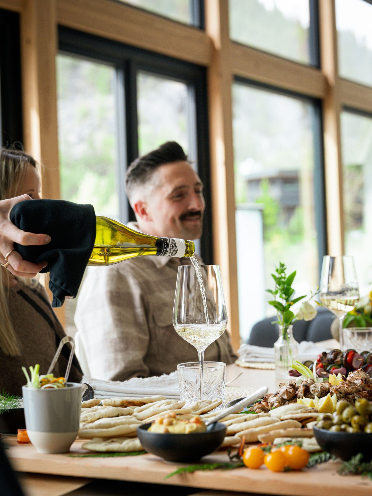 Smiling man having white wine poured at a dining table with charcuterie spread.