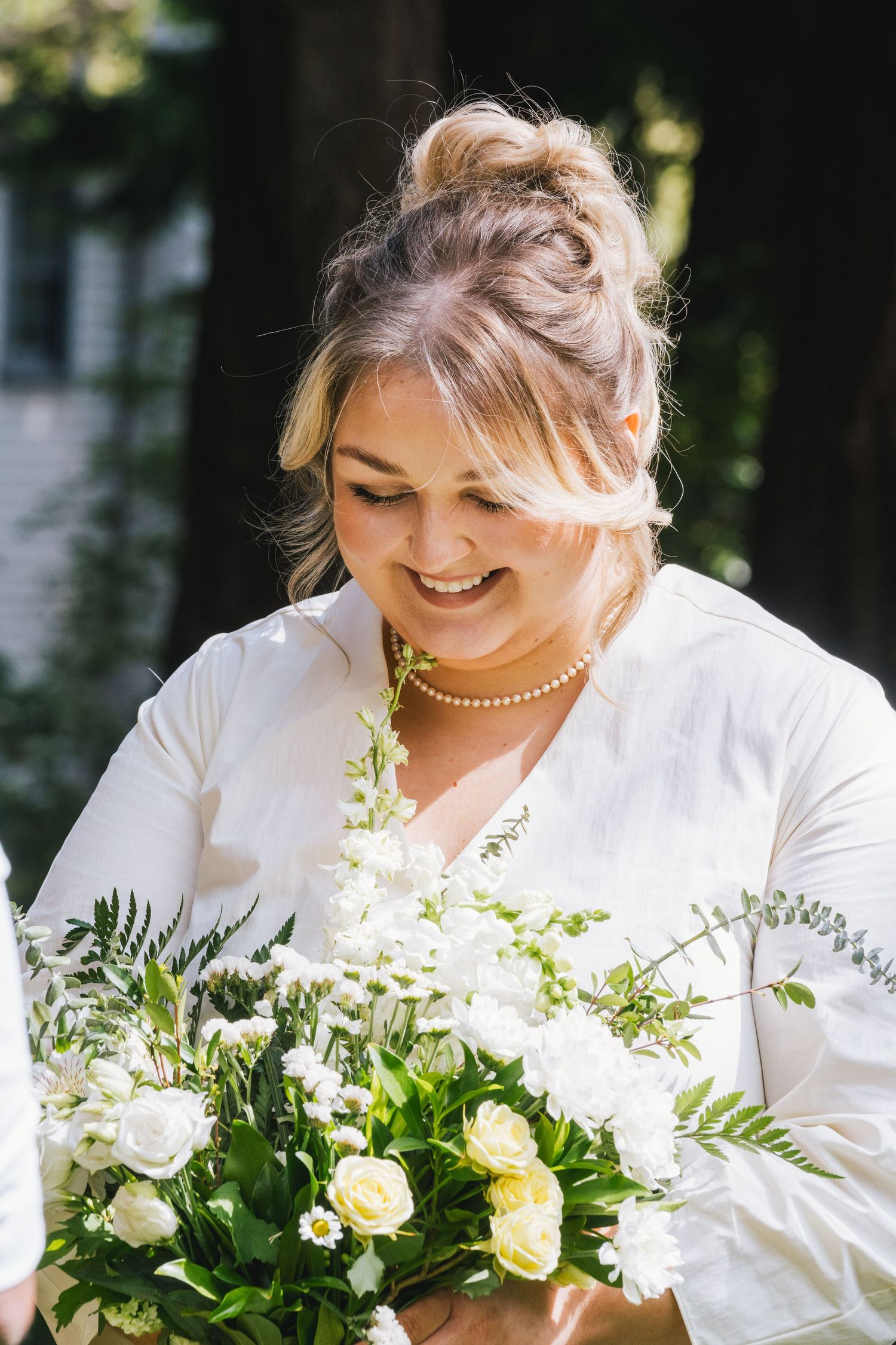 Smiling blonde bride in white dress holding elegant bouquet of white roses and greenery outdoors.