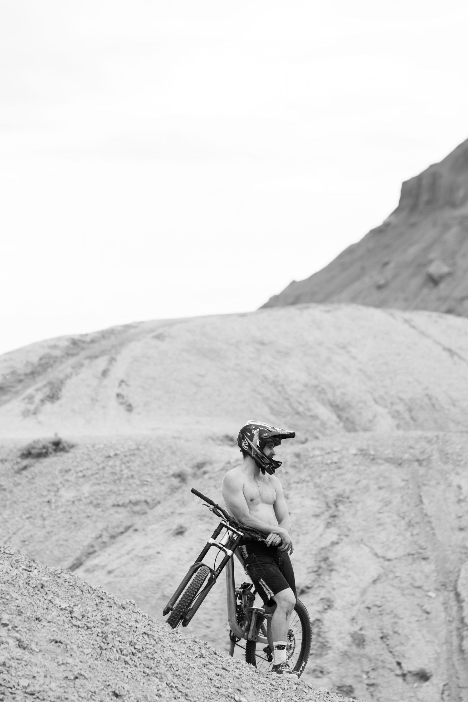 Shirtless mountain biker with helmet resting beside his bike on a rugged desert hillside in black and white.
