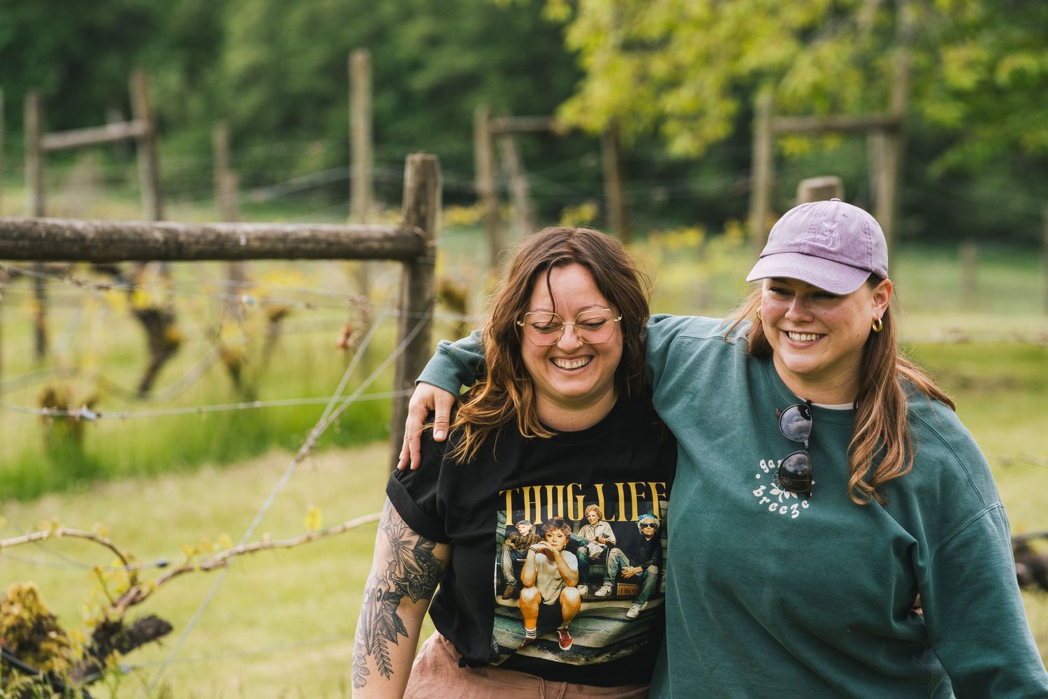 Two smiling women hugging in front of a rustic vineyard fence surrounded by lush green fields.