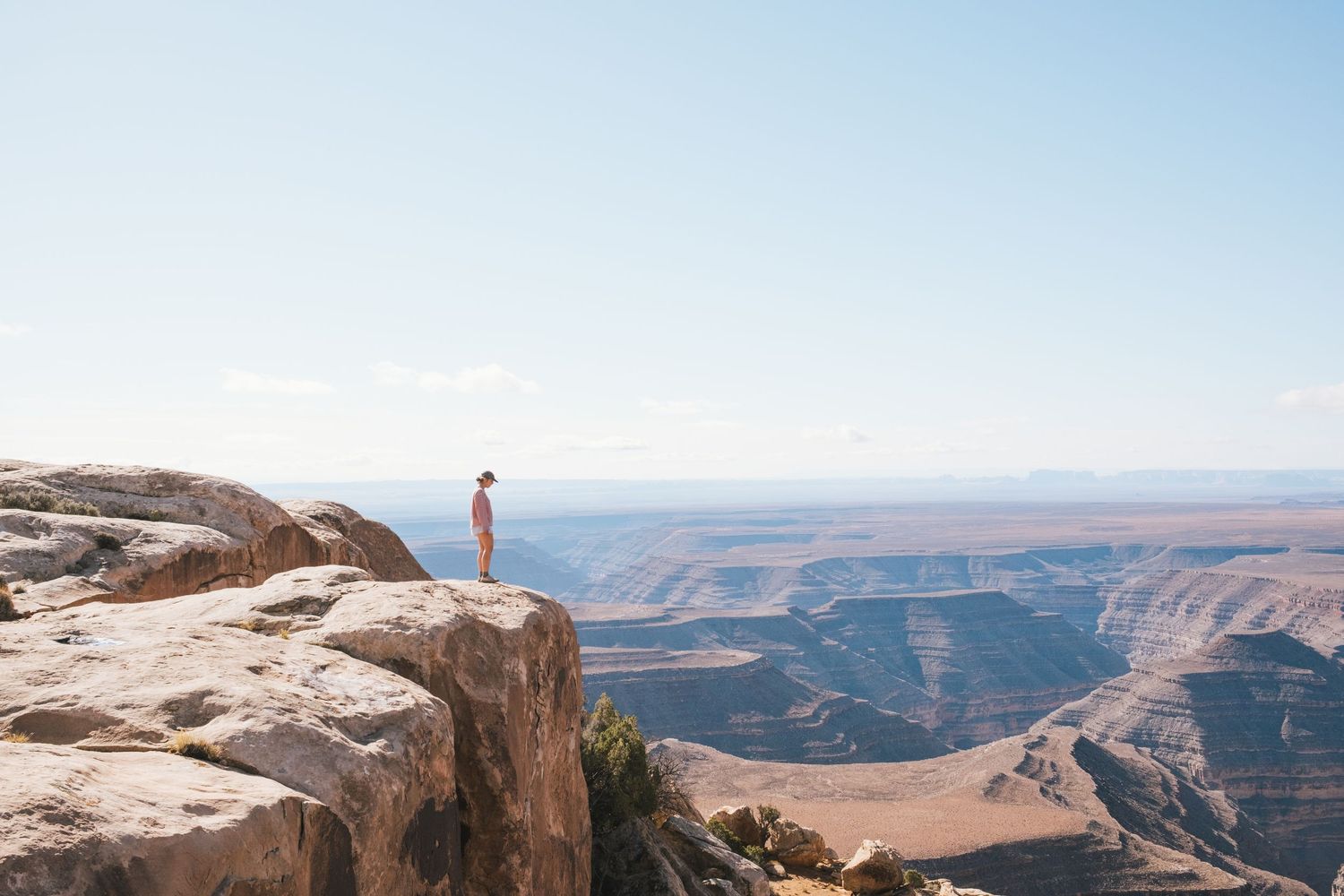 Solo hiker stands on rocky canyon edge overlooking vast desert landscape under clear blue sky.