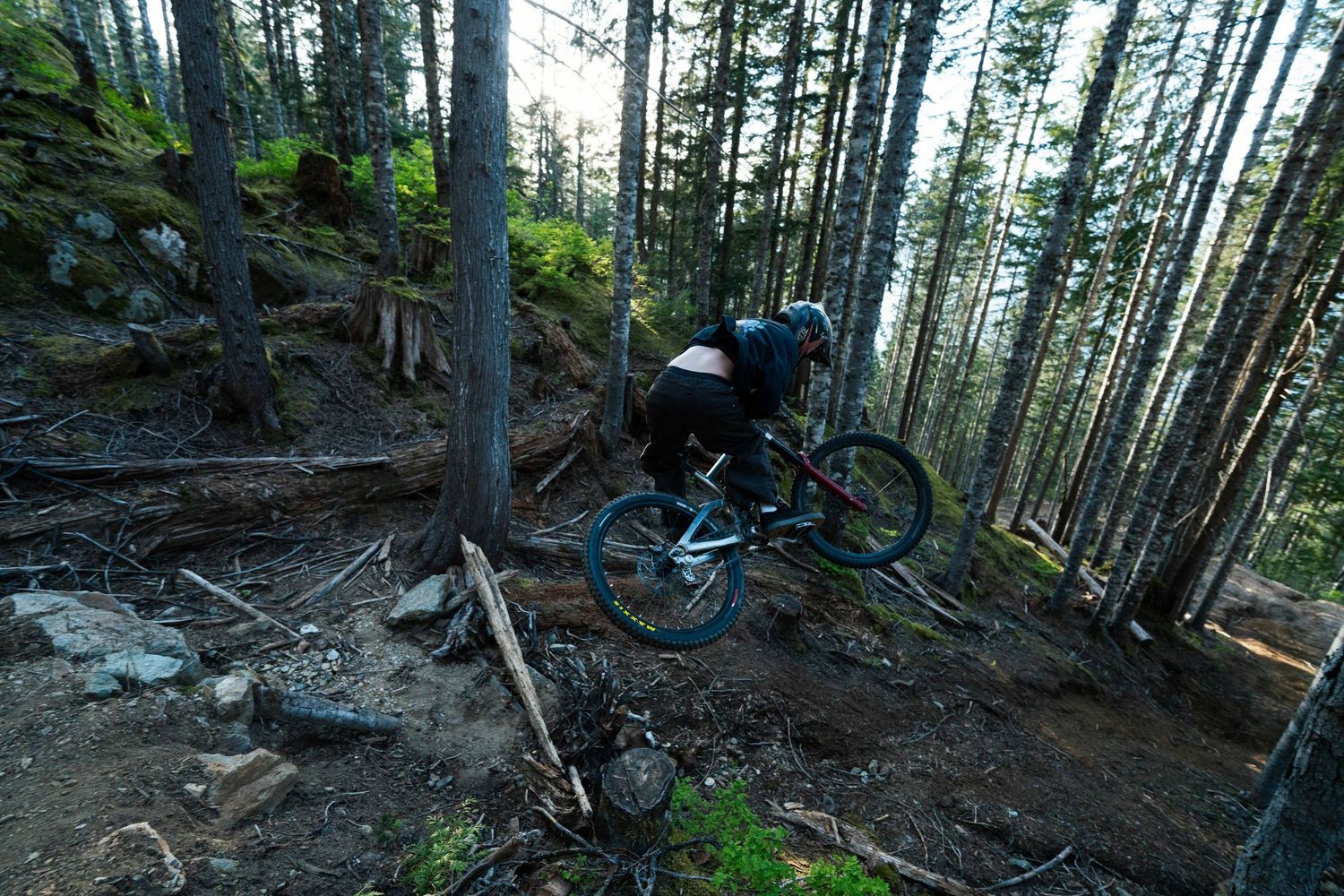 A mountain biker navigates a steep forested trail, leaning into a sharp turn among tall trees.
