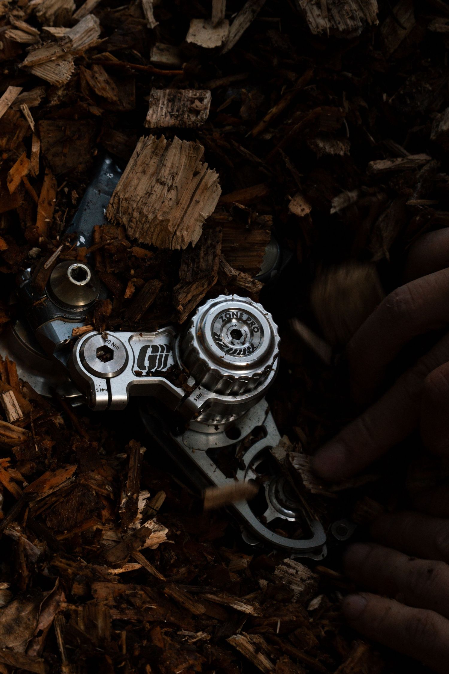 Close-up of a dirty derailleur and bike components resting on a pile of wood chips in dim light.