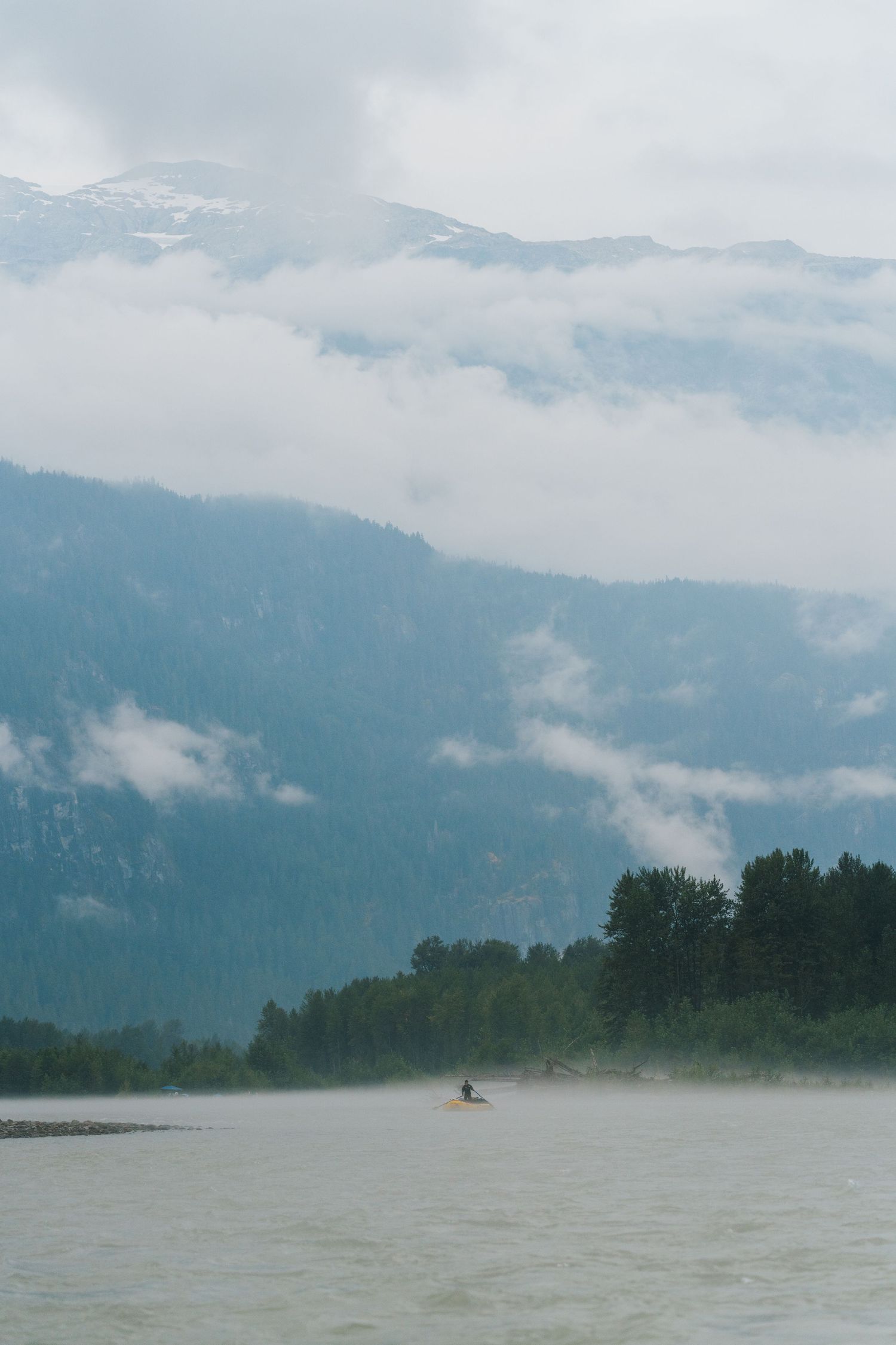 Solo kayaker paddling on a misty mountain lake surrounded by cloud-covered peaks and evergreen forests.