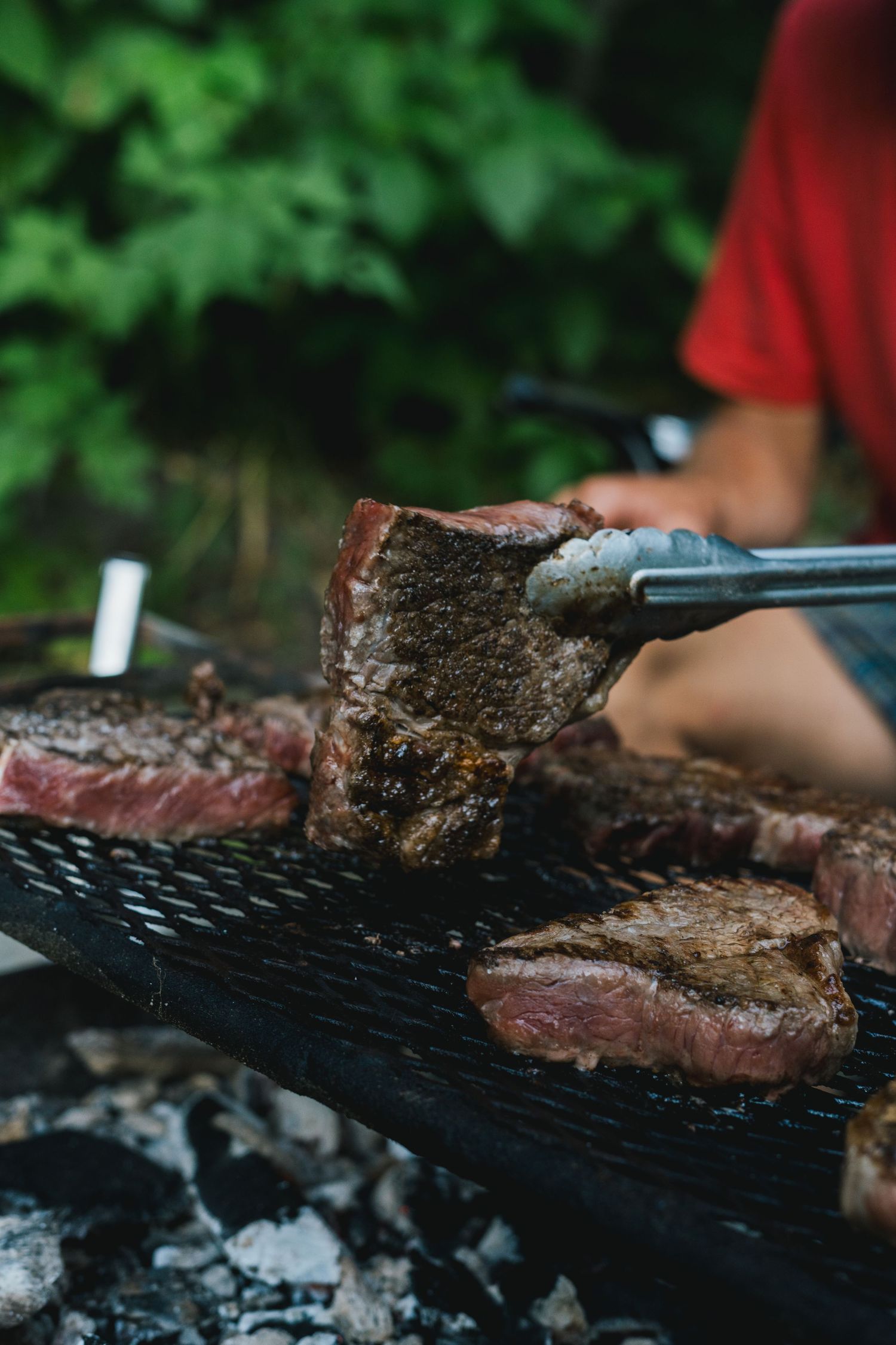 Juicy grilled steaks being turned with tongs on an outdoor charcoal grill surrounded by lush greenery.