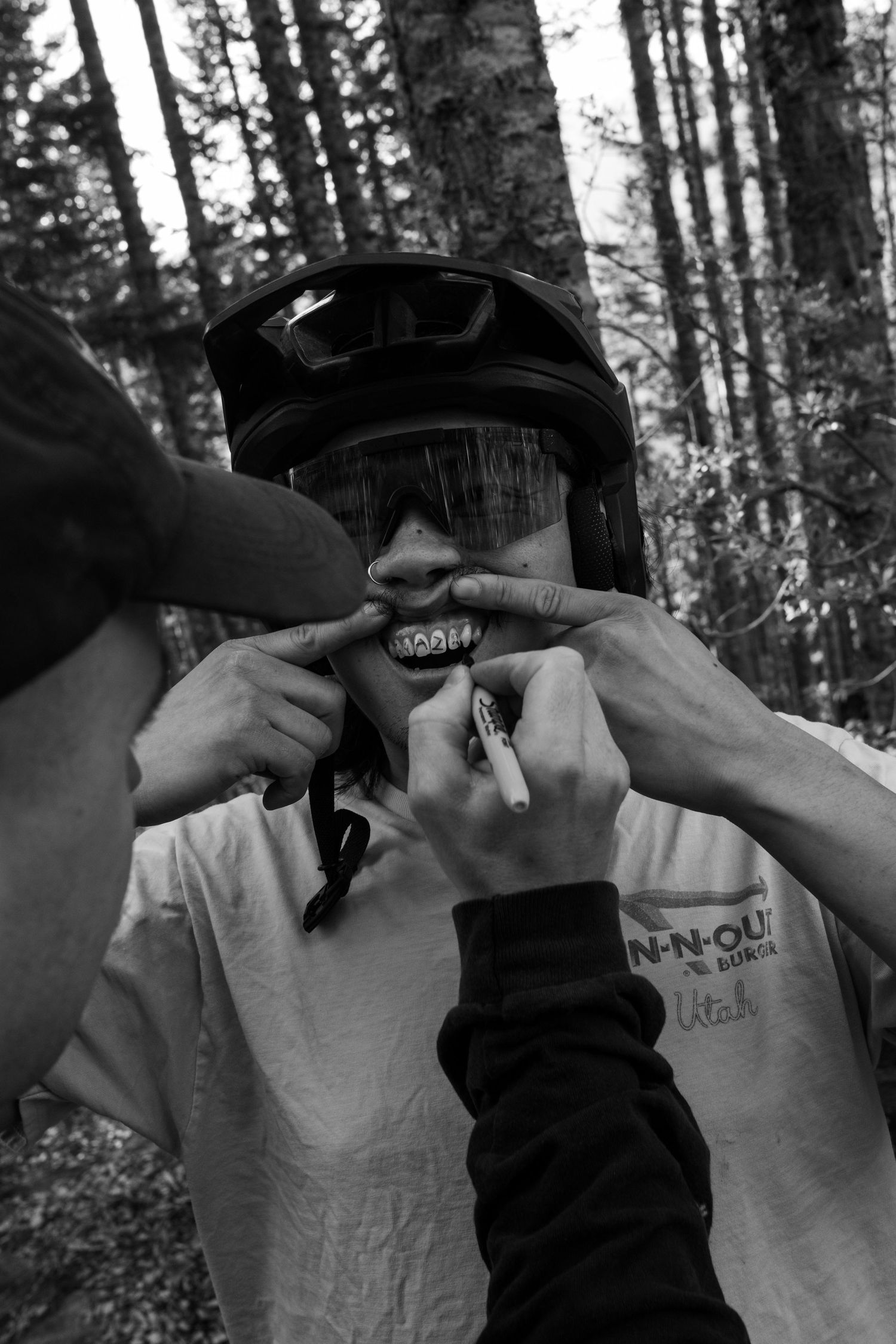Black and white photo of friends drawing a mustache on a helmeted mountain biker in the forest.