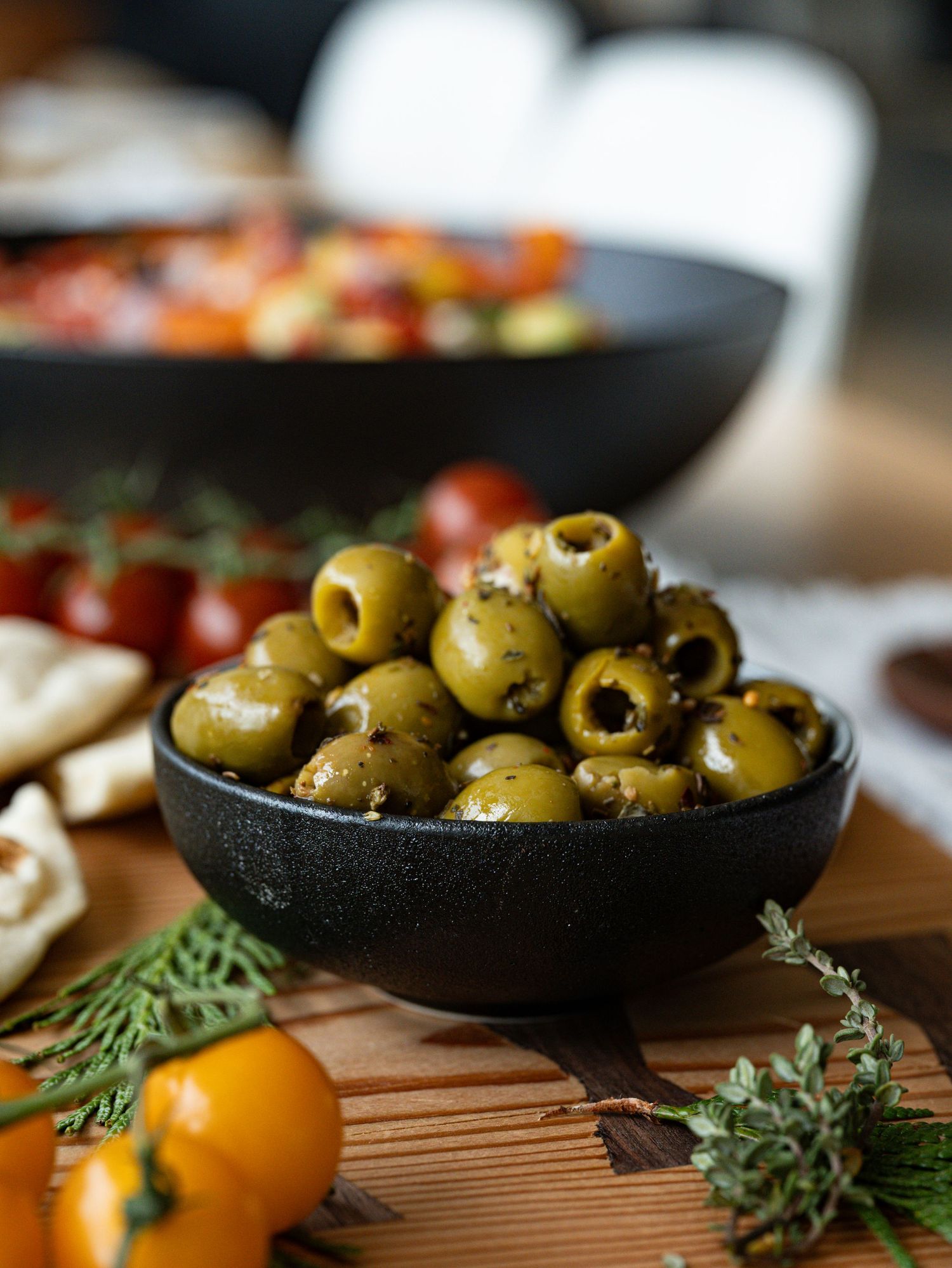 Fresh green olives in a black bowl with cherry tomatoes and herbs on a wooden board.