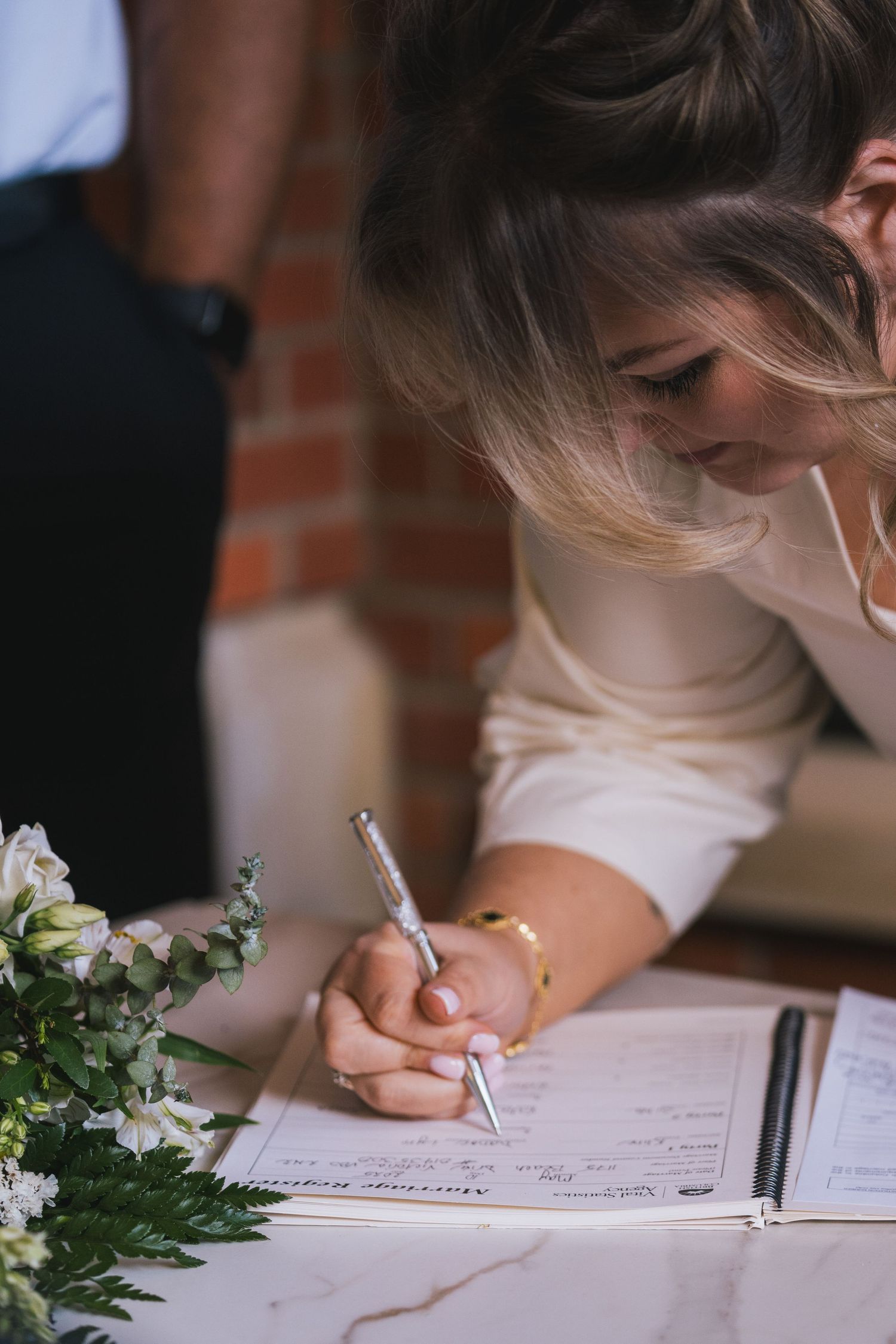 Bride signing marriage register document with pen beside white floral bouquet at ceremony table.