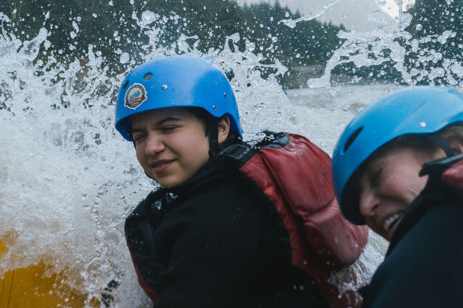 Two people in blue helmets and wetsuits navigating white water rapids while rafting.