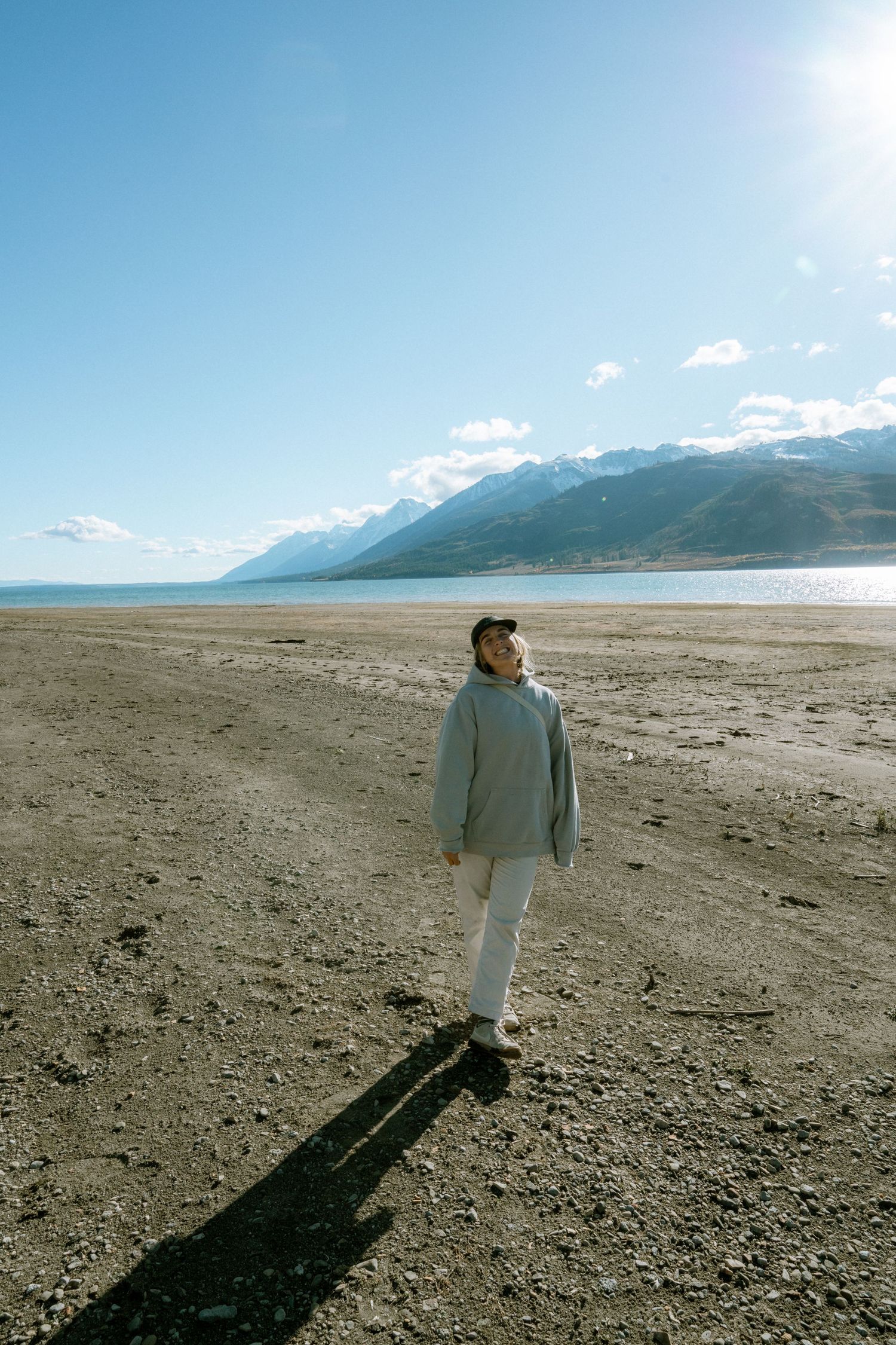 Person in grey hoodie standing on vast sandy beach with snow-capped mountains and blue sky.