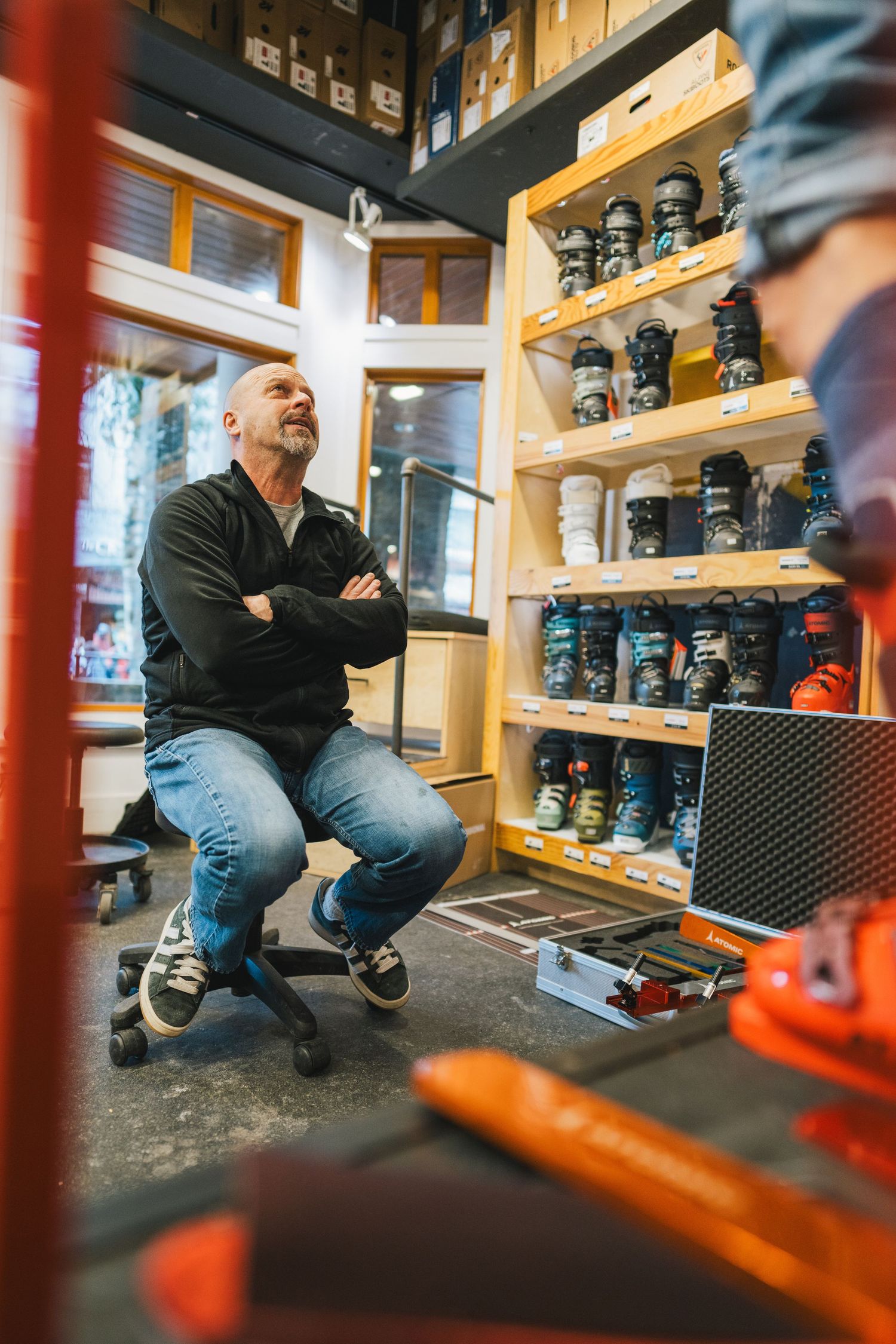 Bald man sitting with arms crossed in a ski equipment rental shop with boots on shelves.