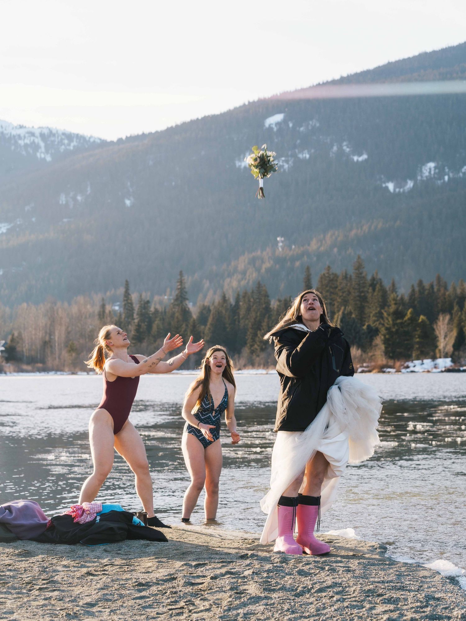 Bride in white skirt and pink boots tosses bouquet to friends in swimsuits by a frozen mountain lake.