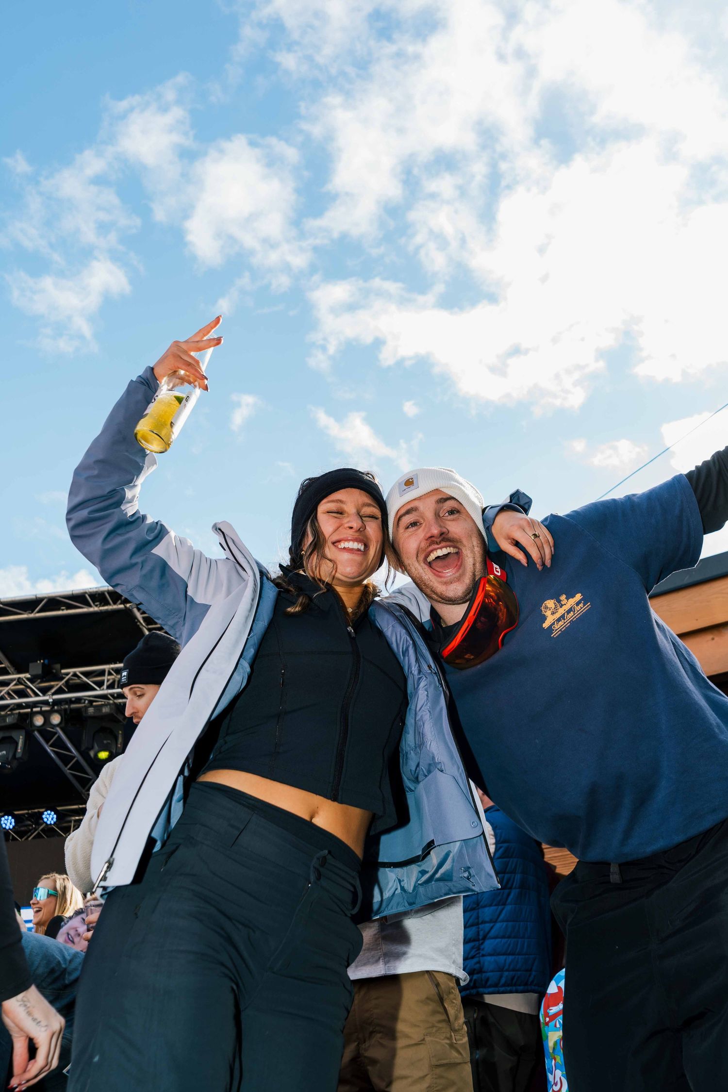 Two friends laughing and celebrating outdoors at a ski resort music festival under blue sky.