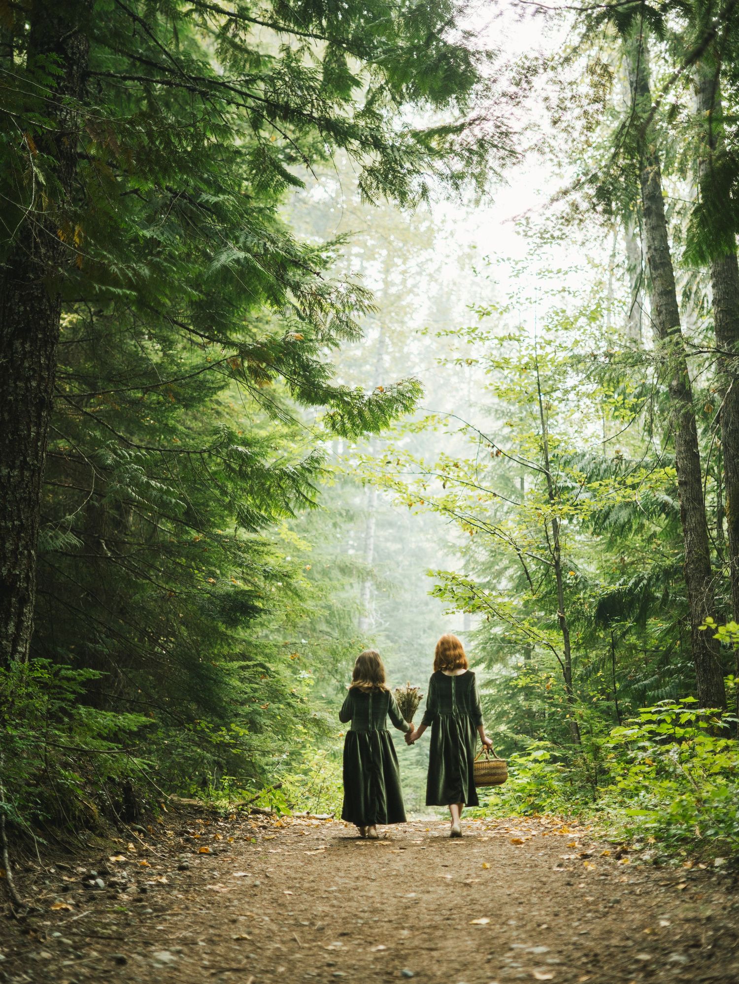 Two red-haired girls in matching dark dresses walking hand in hand along misty forest path.