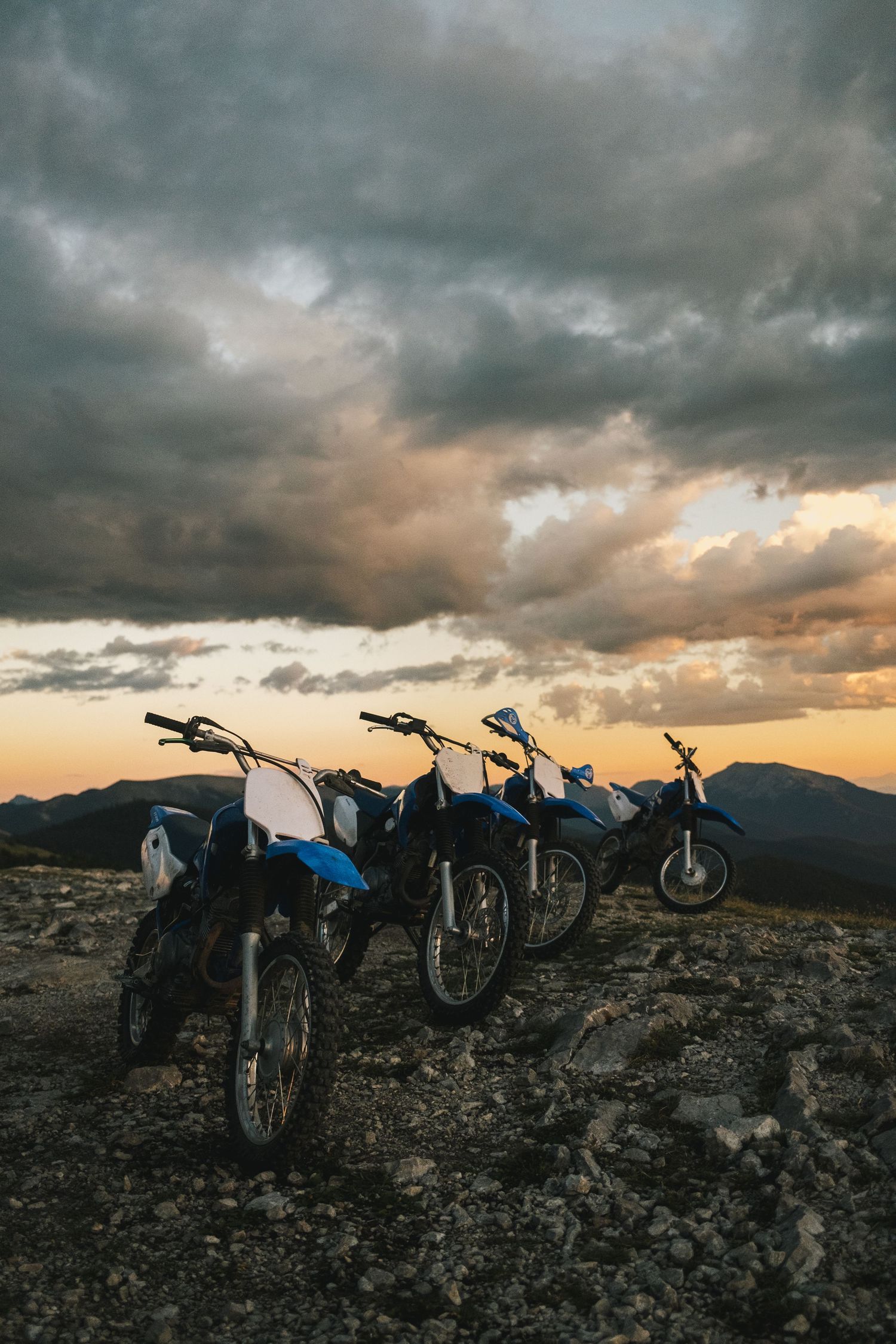 Four dirt bikes parked in a row on rocky terrain under a dramatic sunset sky.