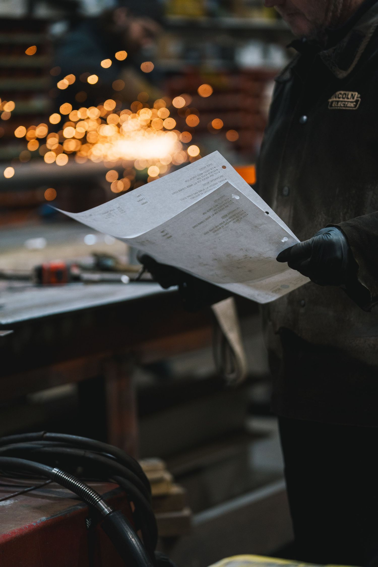 A worker in a Lincoln Electric jacket reviews technical documents with welding sparks in the background.