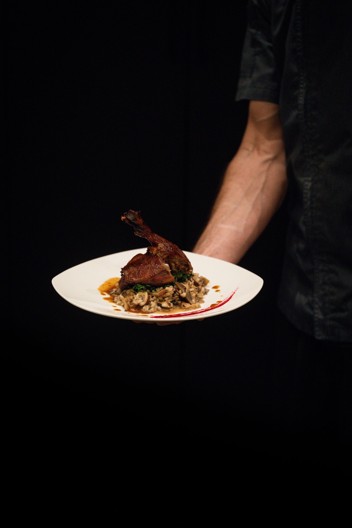Chef presenting a gourmet duck leg dish on white plate with rice on black background.