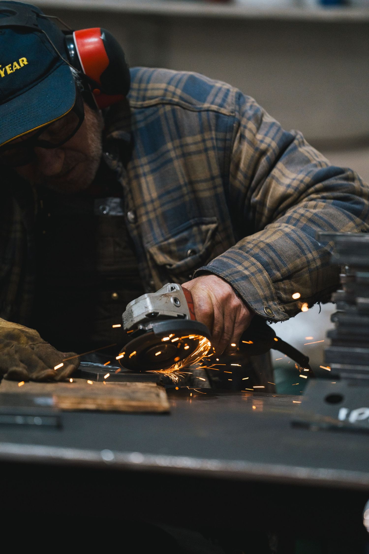 A worker wearing ear protection uses an angle grinder on metal, producing bright sparks in a workshop.