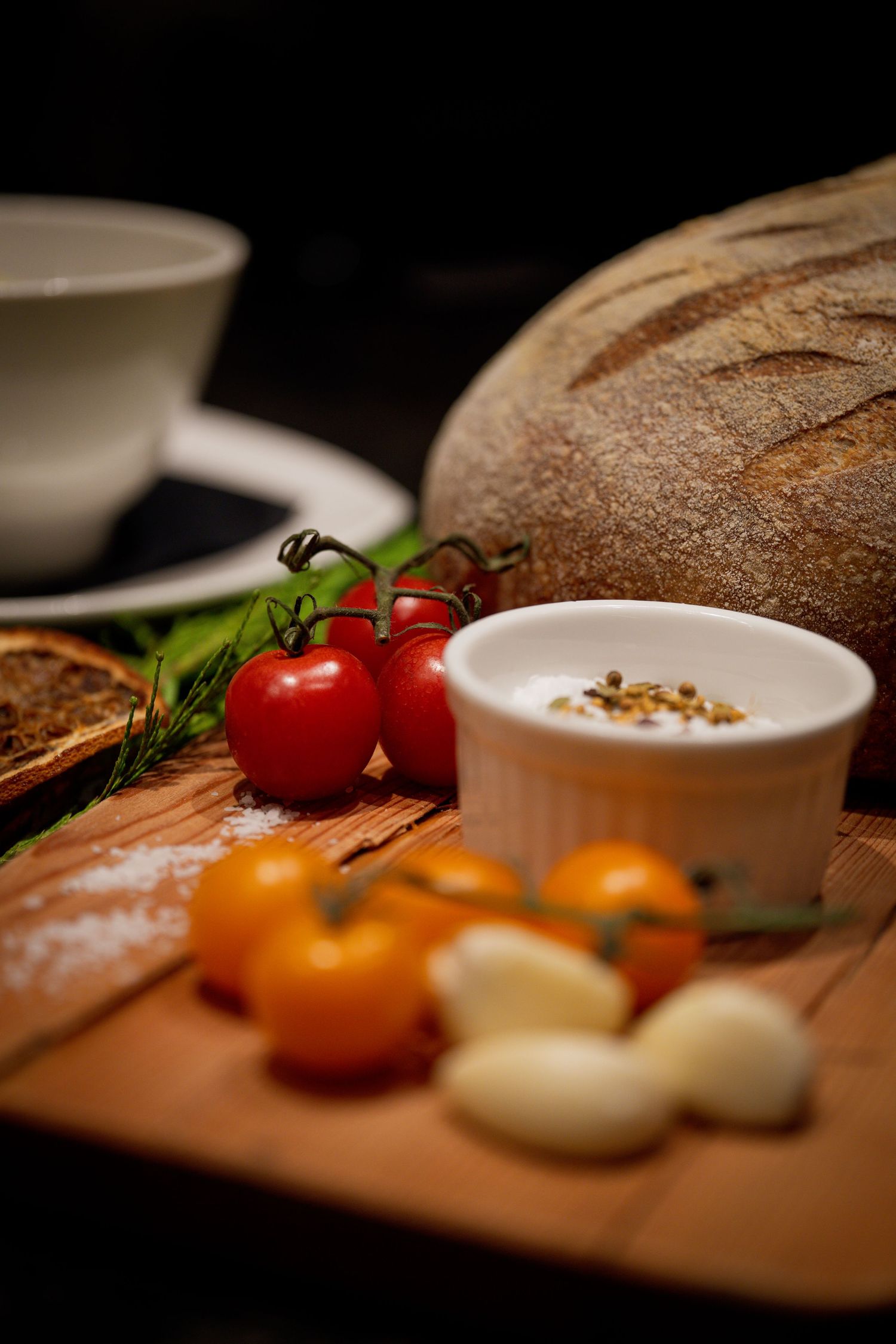 Rustic wooden board with cherry tomatoes, garlic, herbs, dipping sauce, and artisan bread loaf.