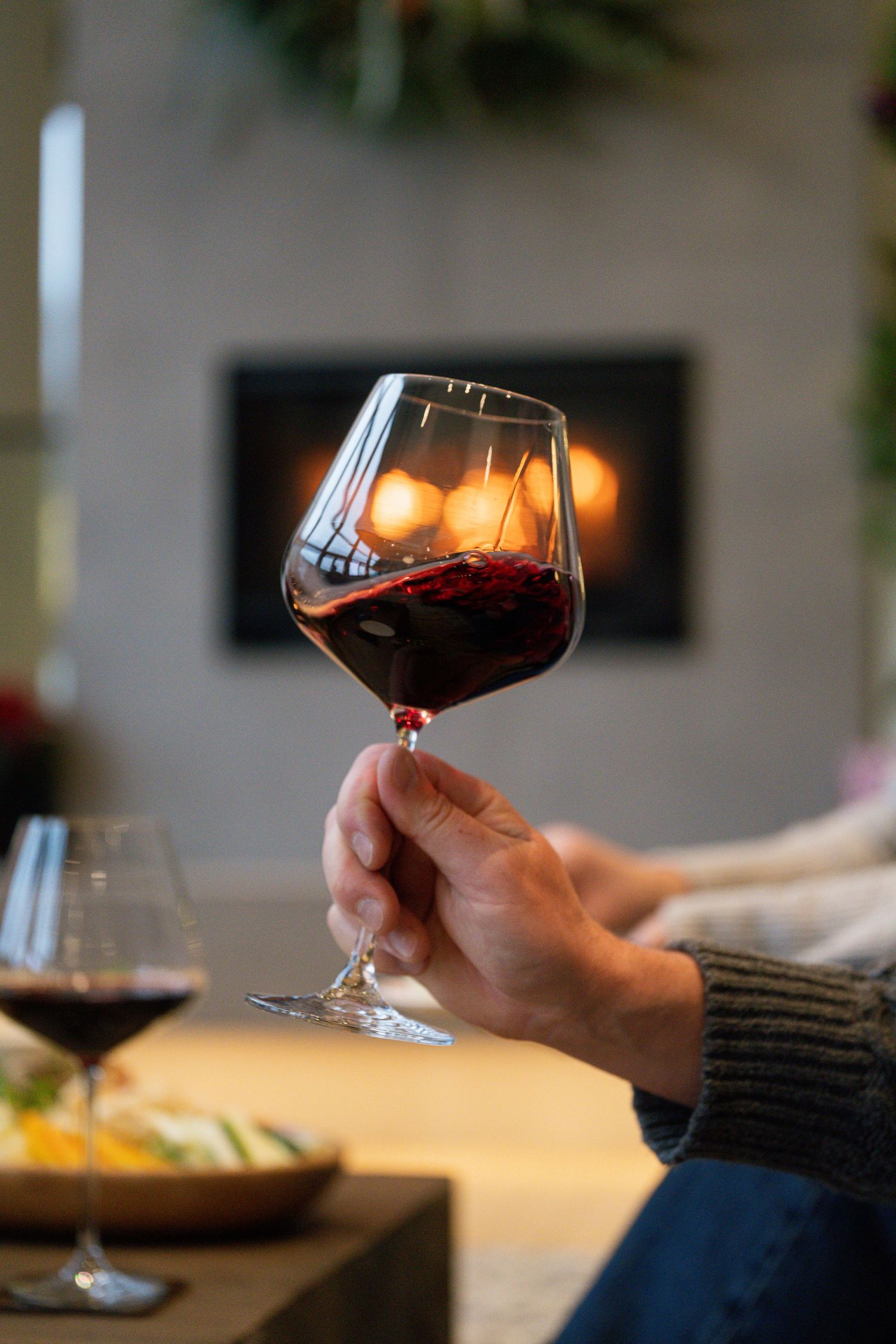 Hand holding a swirling glass of red wine near a glowing fireplace in a cozy living room.