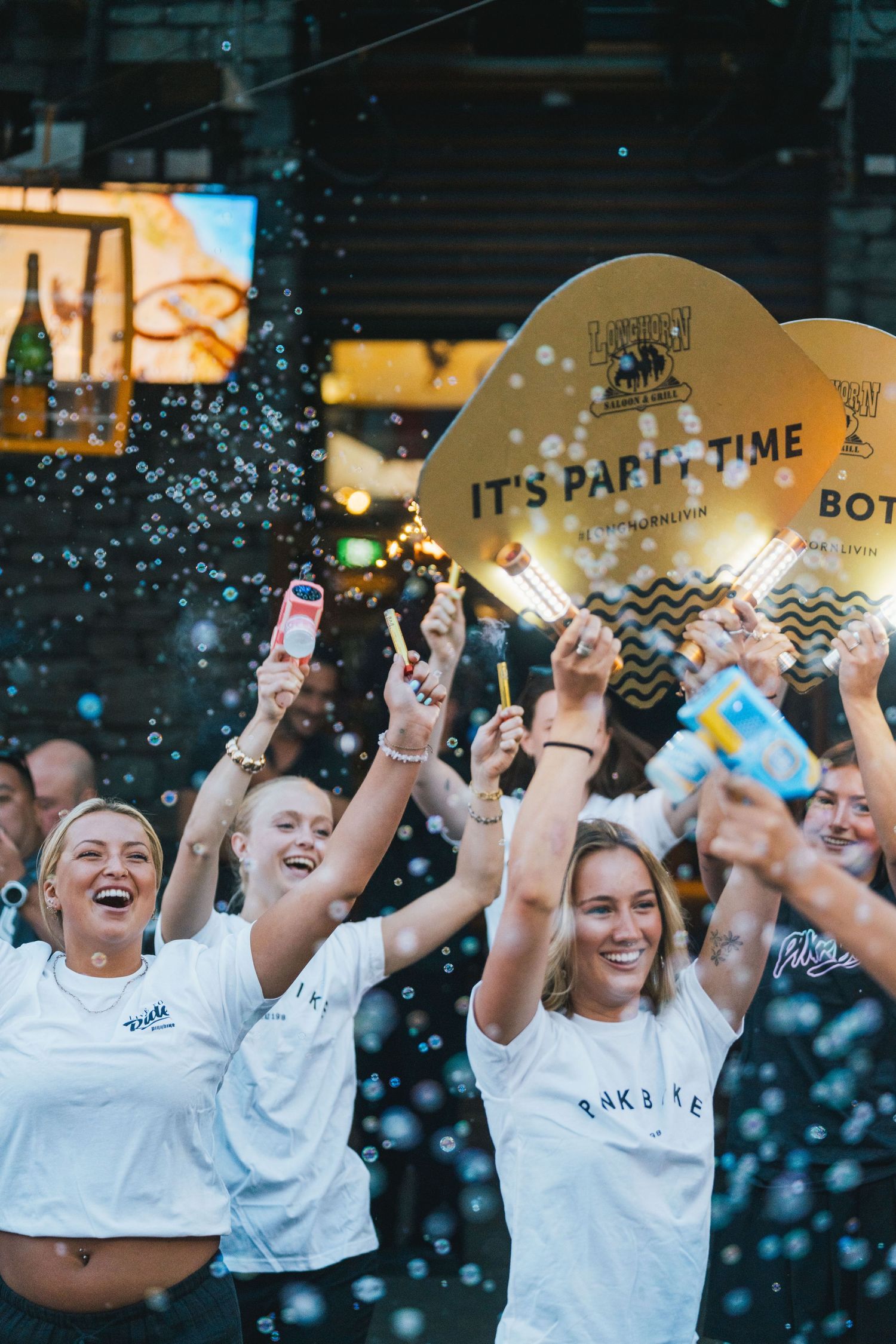 Excited crowd celebrating at an outdoor party event with bubbles and Its Party Time signs.
