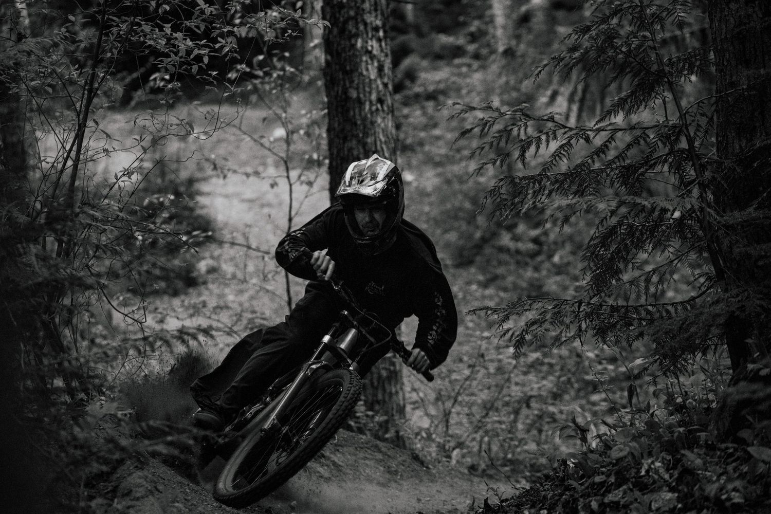 Black and white photo of a mountain biker carving a fast turn through a dense forest trail.