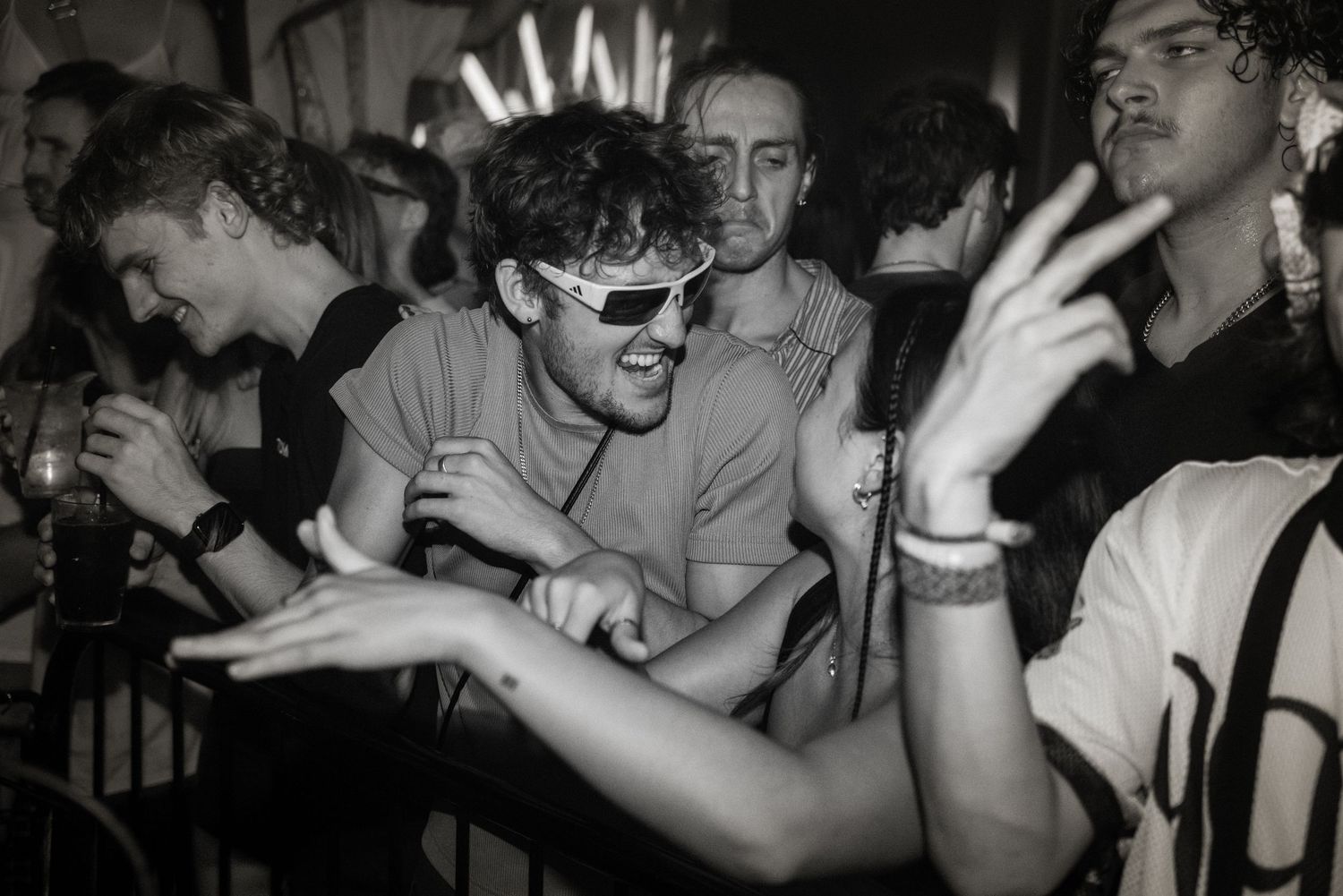 Young man wearing sunglasses dancing energetically at a crowded nightclub party in black and white photo.
