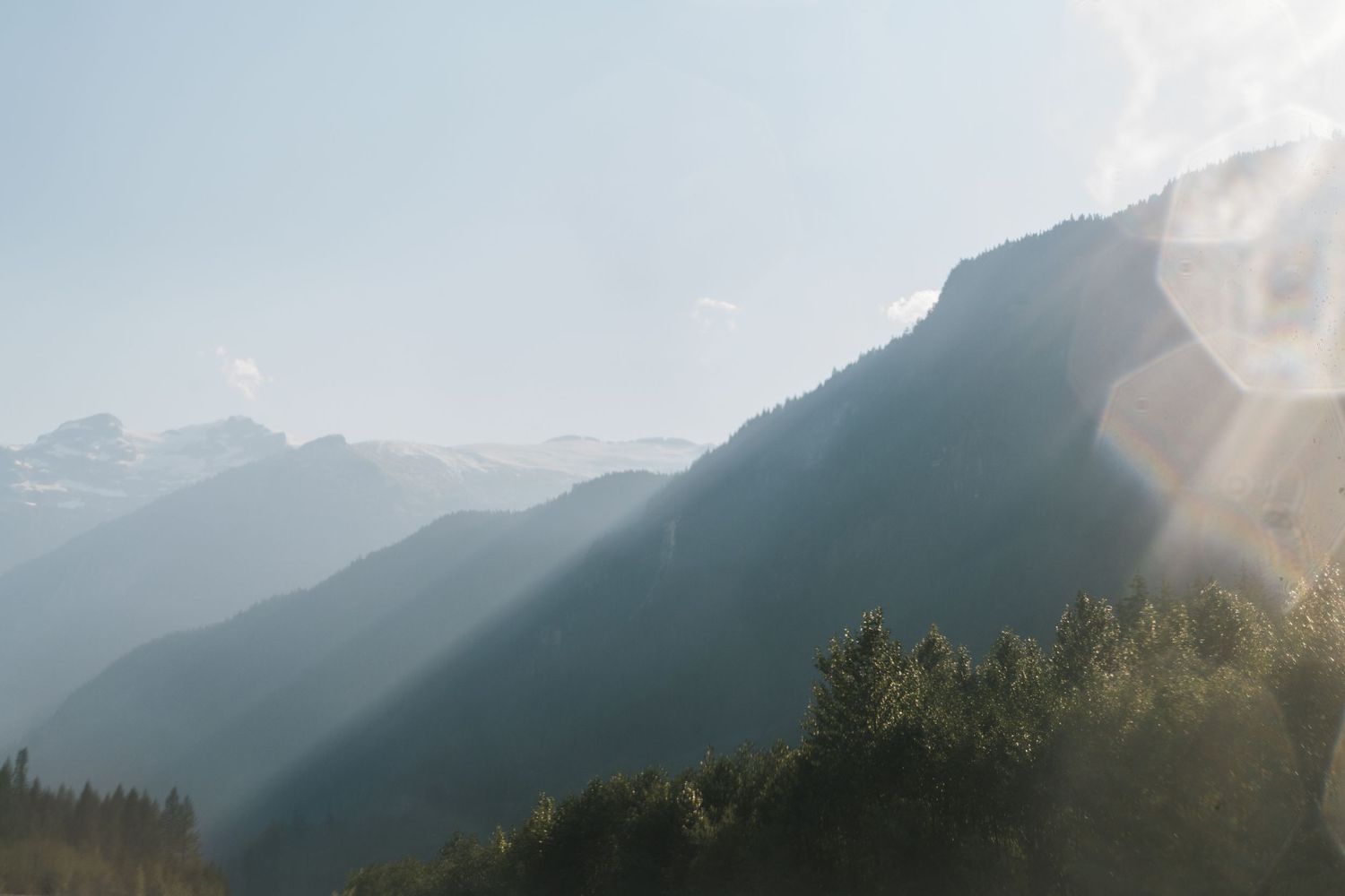 Misty mountain landscape with sunbeams filtering through hazy skies over forested alpine peaks.