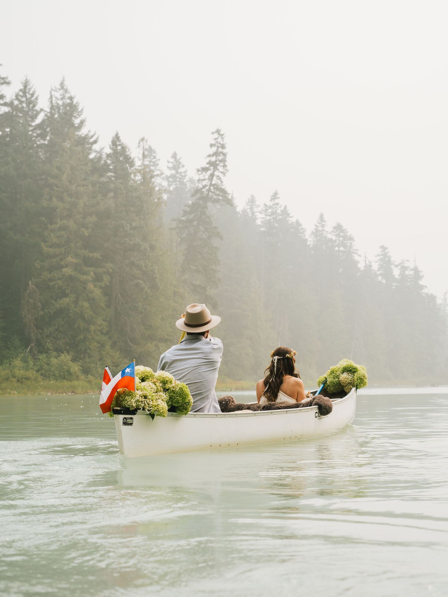 Couple canoeing on a misty lake surrounded by evergreen forest with flowers and a flag in the boat.