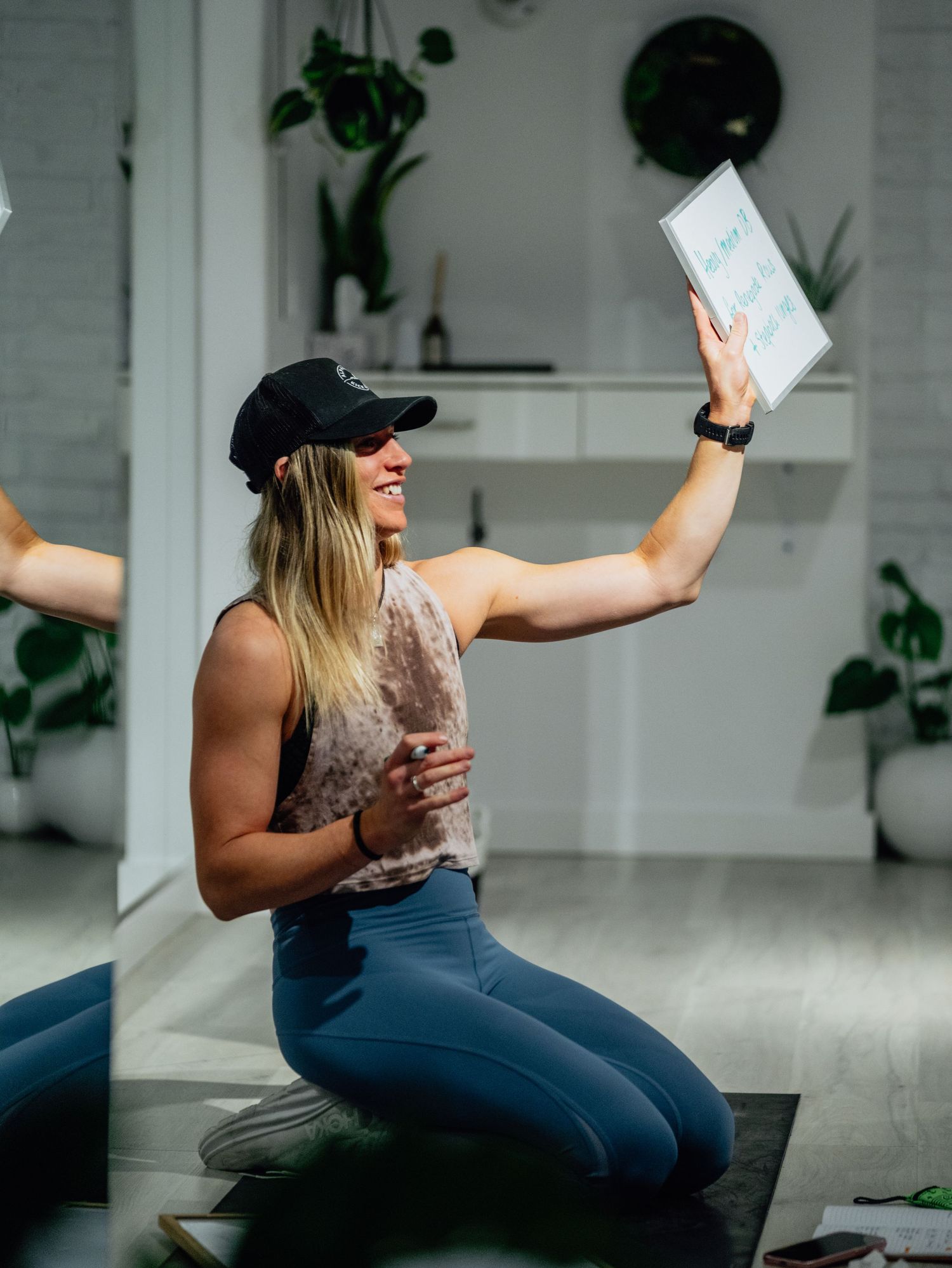 Smiling woman in cap and leggings kneeling on yoga mat holding a framed sign indoors.