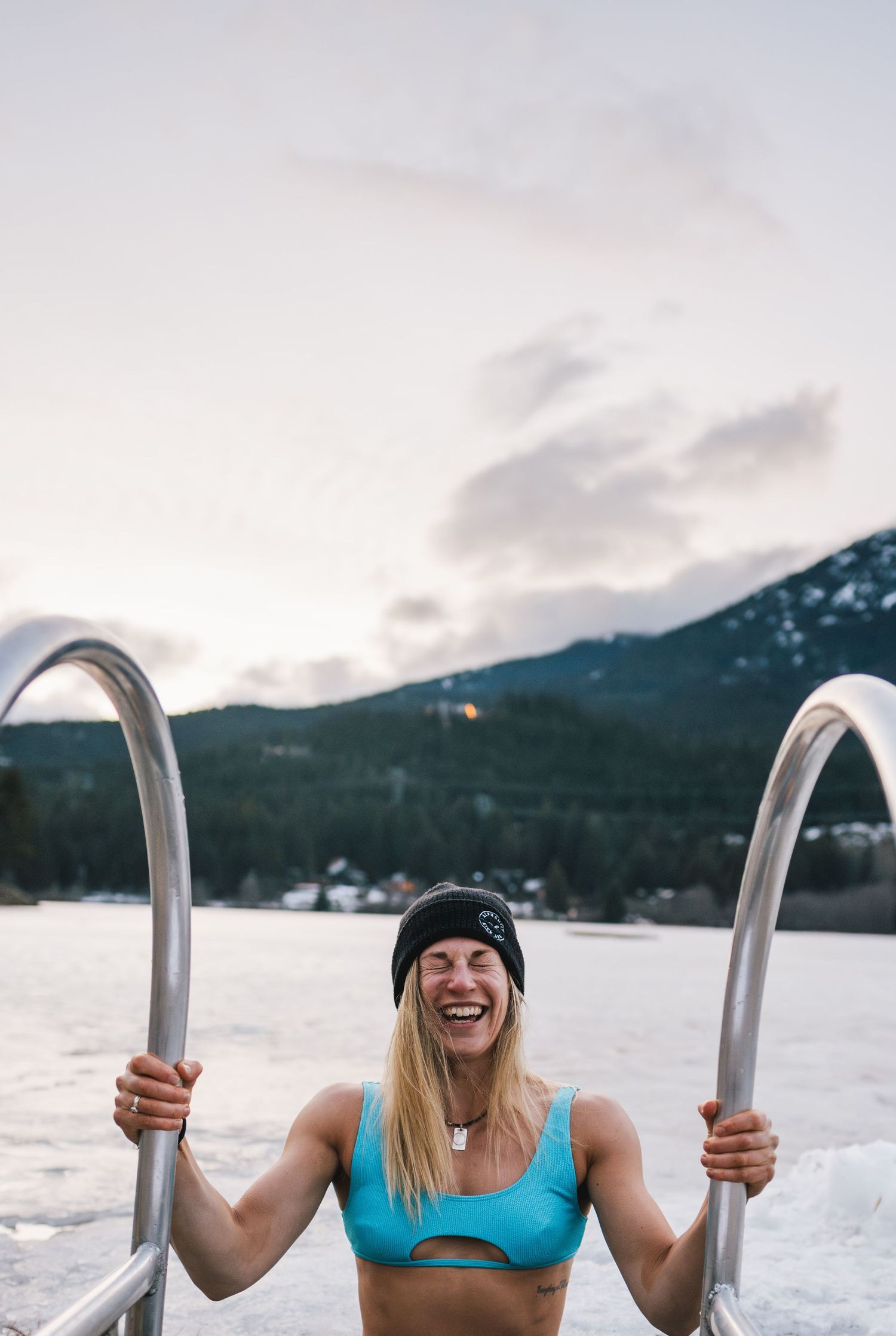 Smiling woman in a blue bikini top and black beanie emerging from an icy lake in winter.