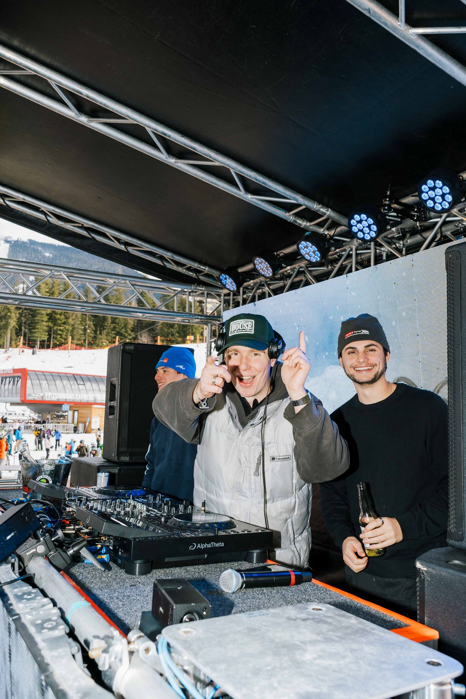 Two DJs performing at an outdoor ski resort stage with DJ equipment and mountain backdrop.