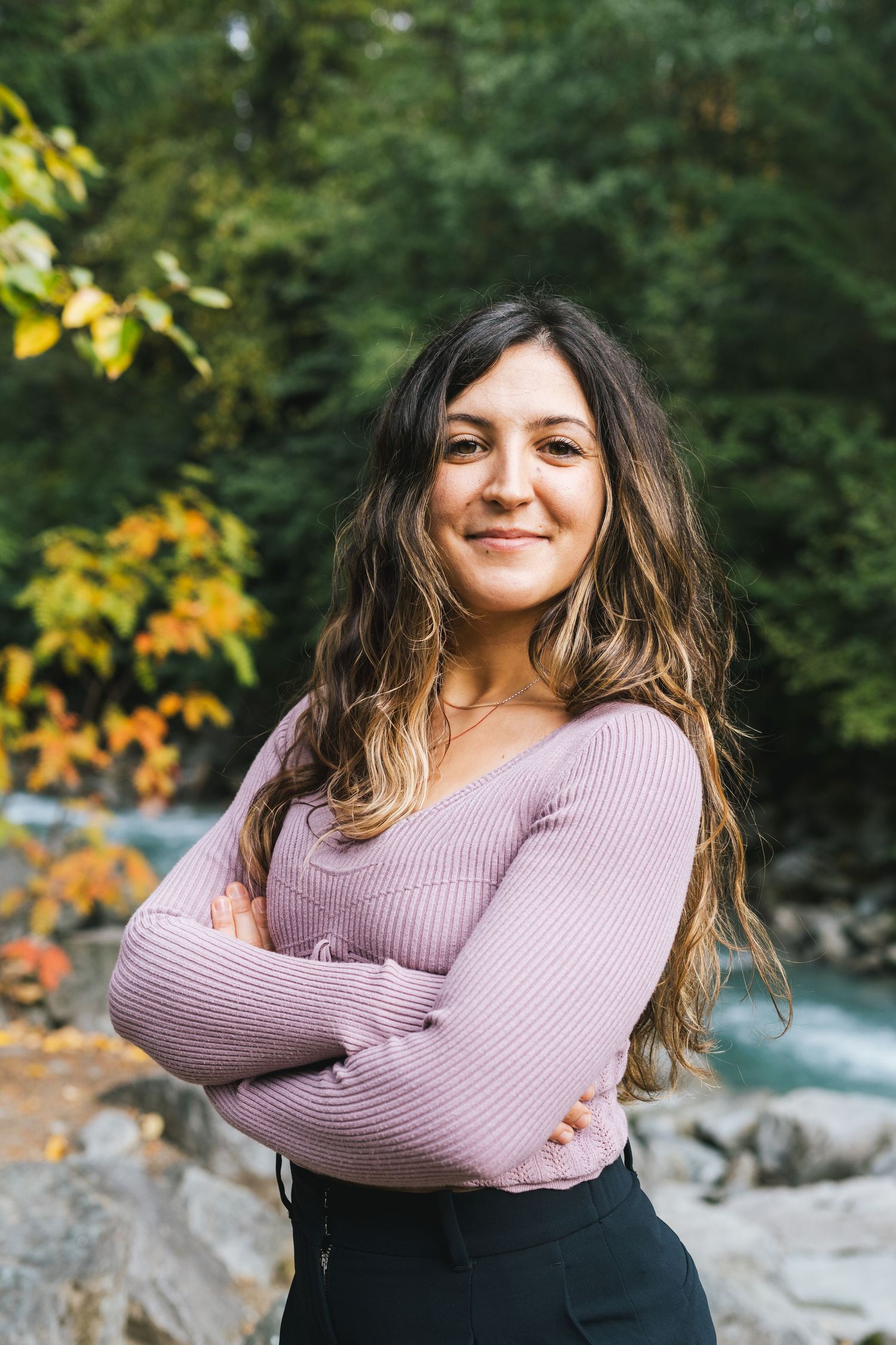Smiling brunette woman with arms crossed standing outdoors near a river in autumn forest.
