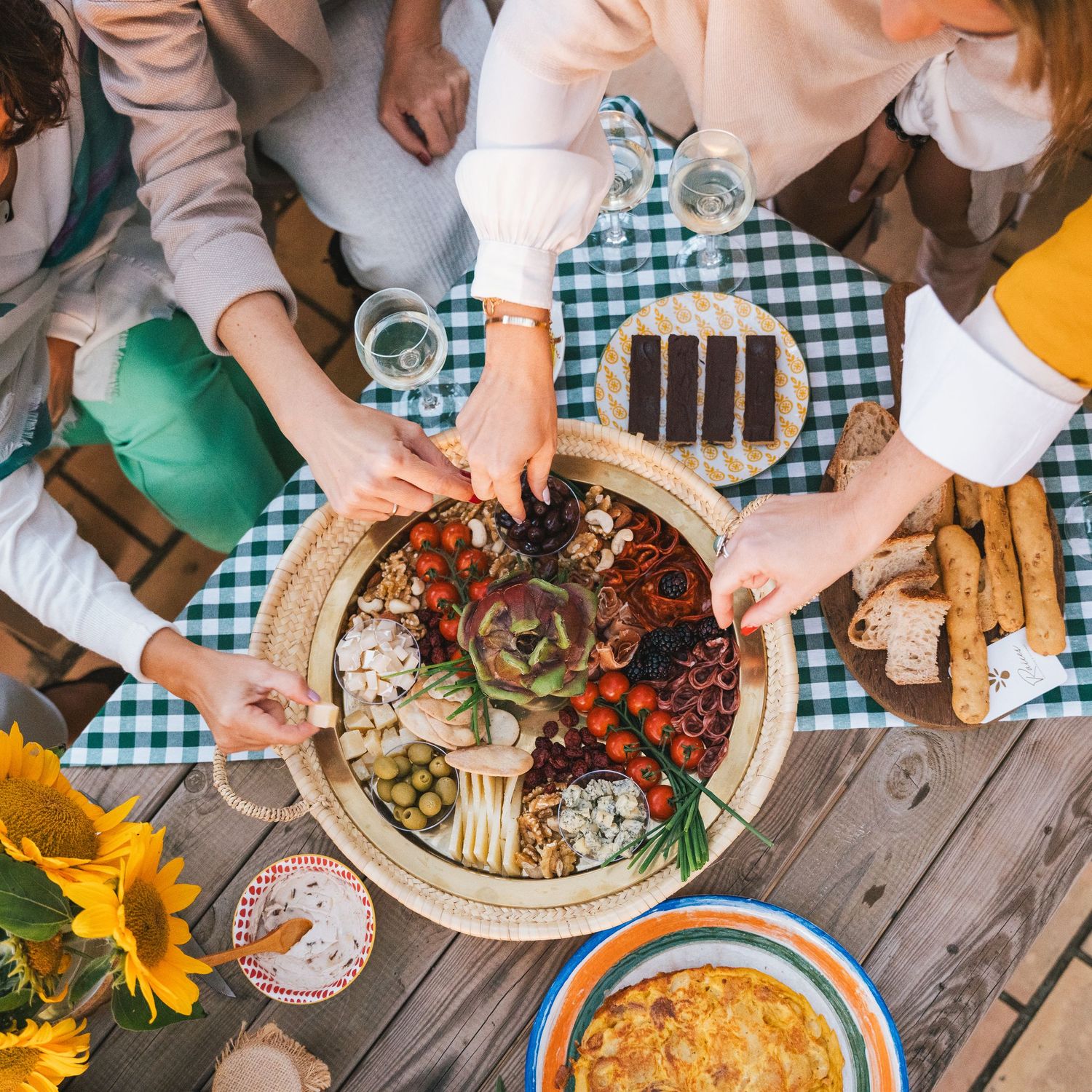 Overhead view of friends sharing a charcuterie board with cheese, vegetables and bread at a picnic table.