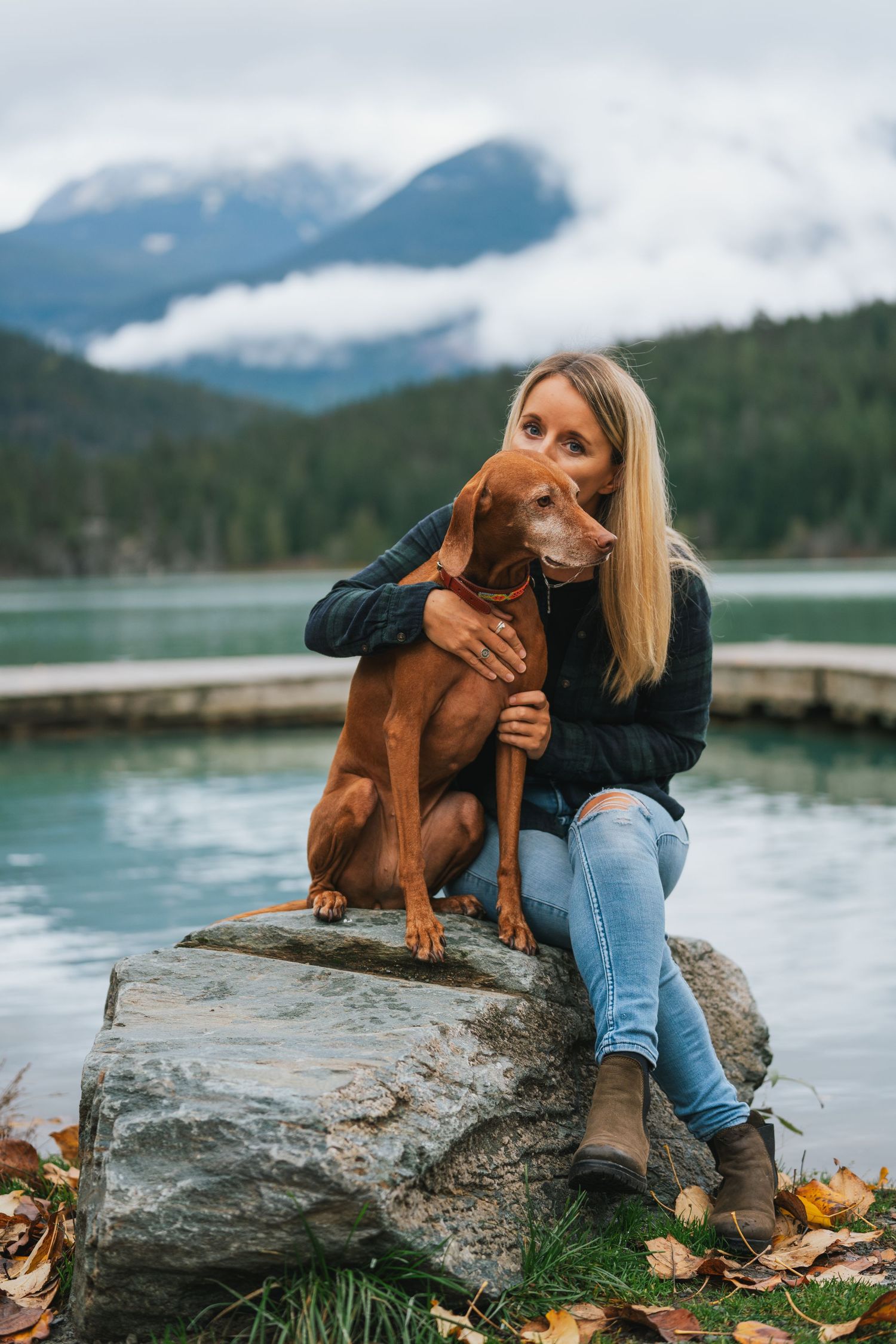 Blonde woman hugging a brown Vizsla dog while sitting on a rock by a mountain lake.