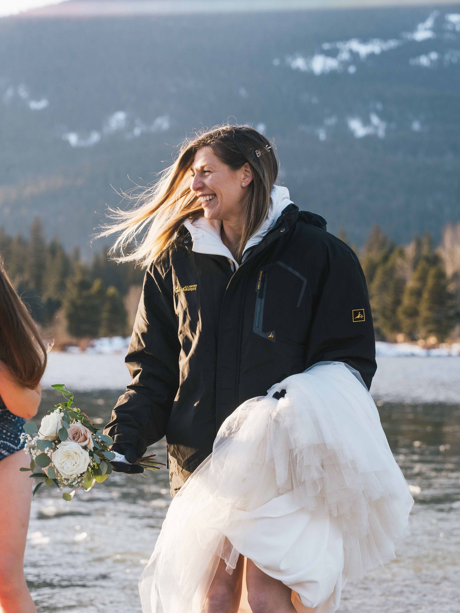 Bride laughing in black jacket holding wedding dress and bouquet at snowy mountain elopement.