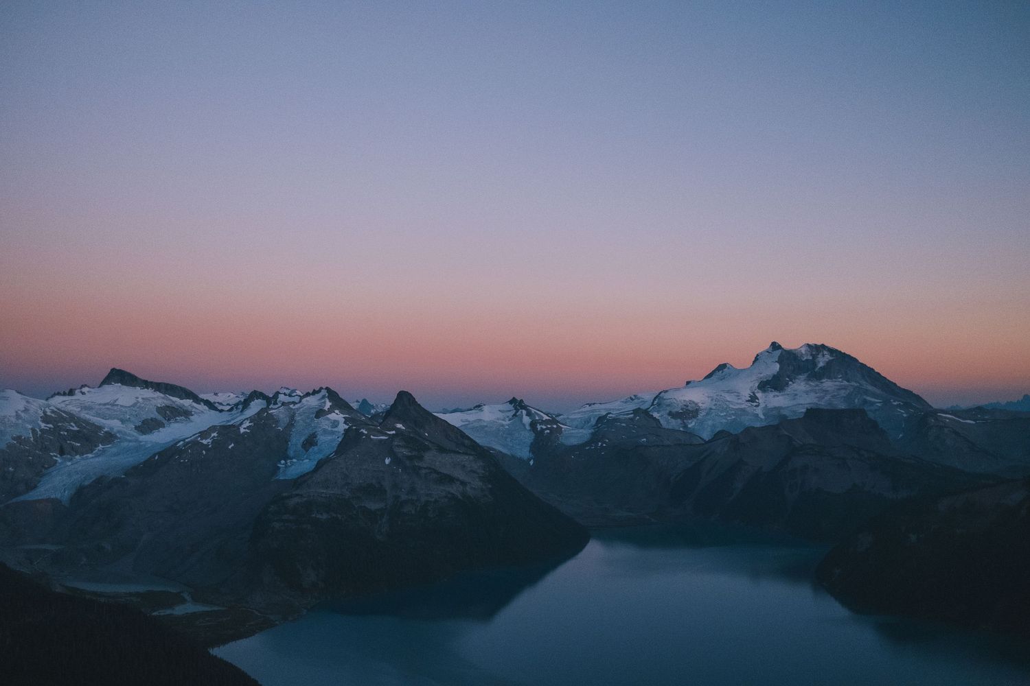 A breathtaking alpine sunset casts pink and purple hues over snow-capped mountain peaks above a serene lake.