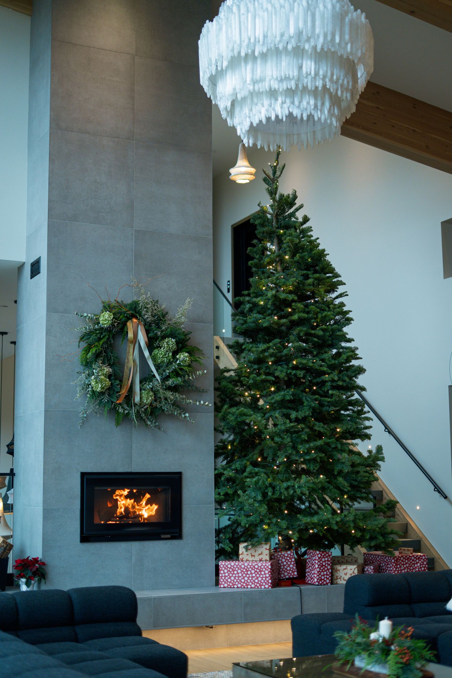 Elegant living room with tall decorated Christmas tree, lit fireplace, and modern chandelier.