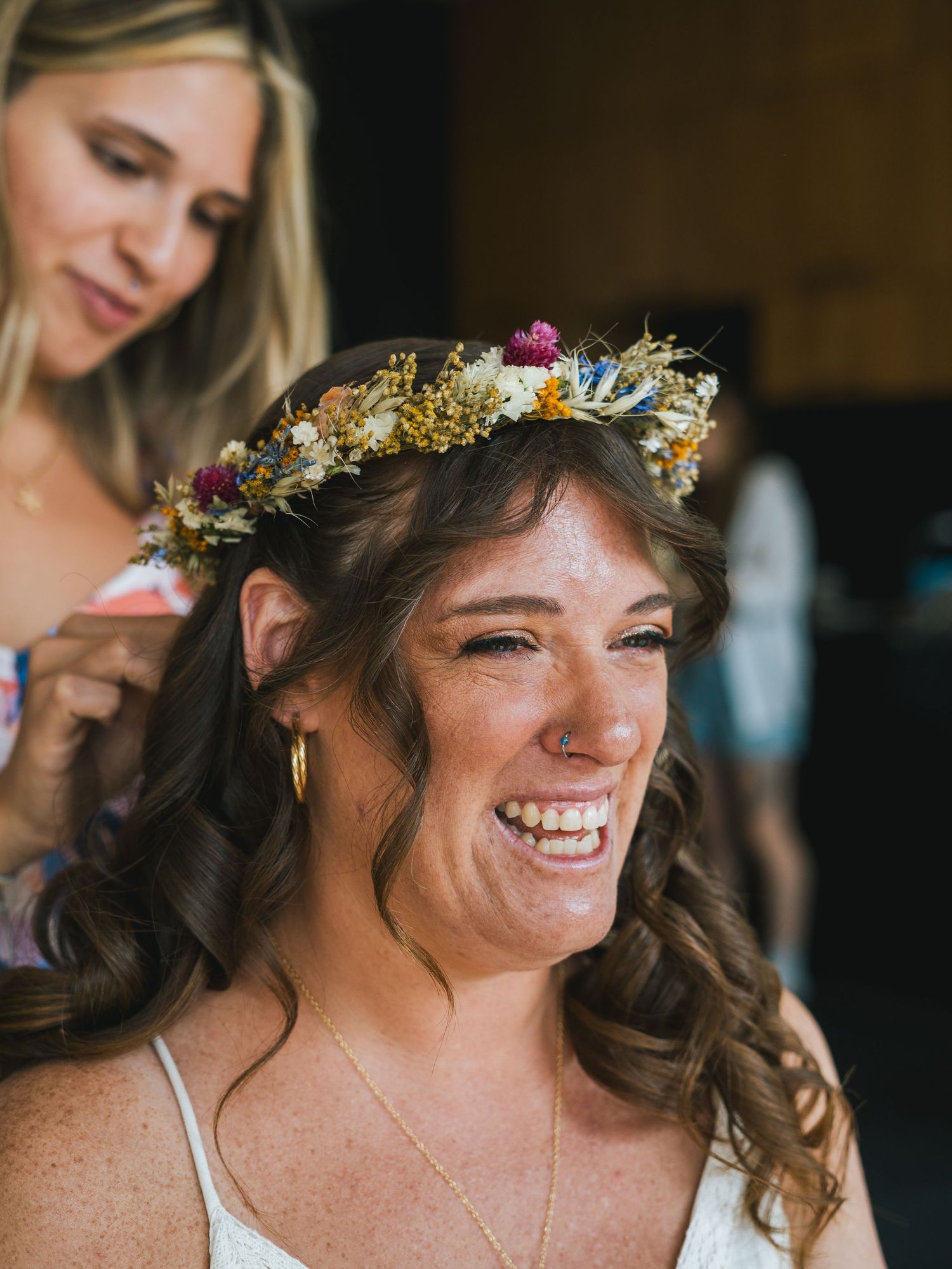 Smiling bride wearing colorful dried flower crown being adjusted by bridesmaid before wedding ceremony.