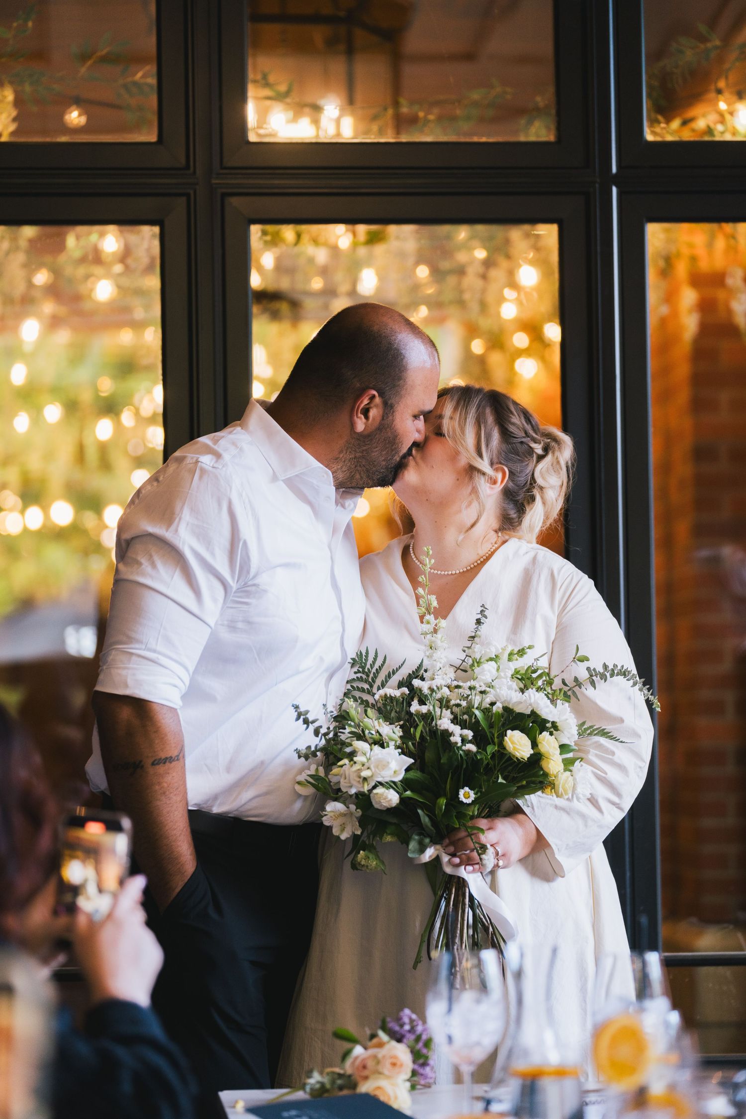 Newlywed couple sharing first kiss holding white floral bouquet before warm fairy light backdrop.