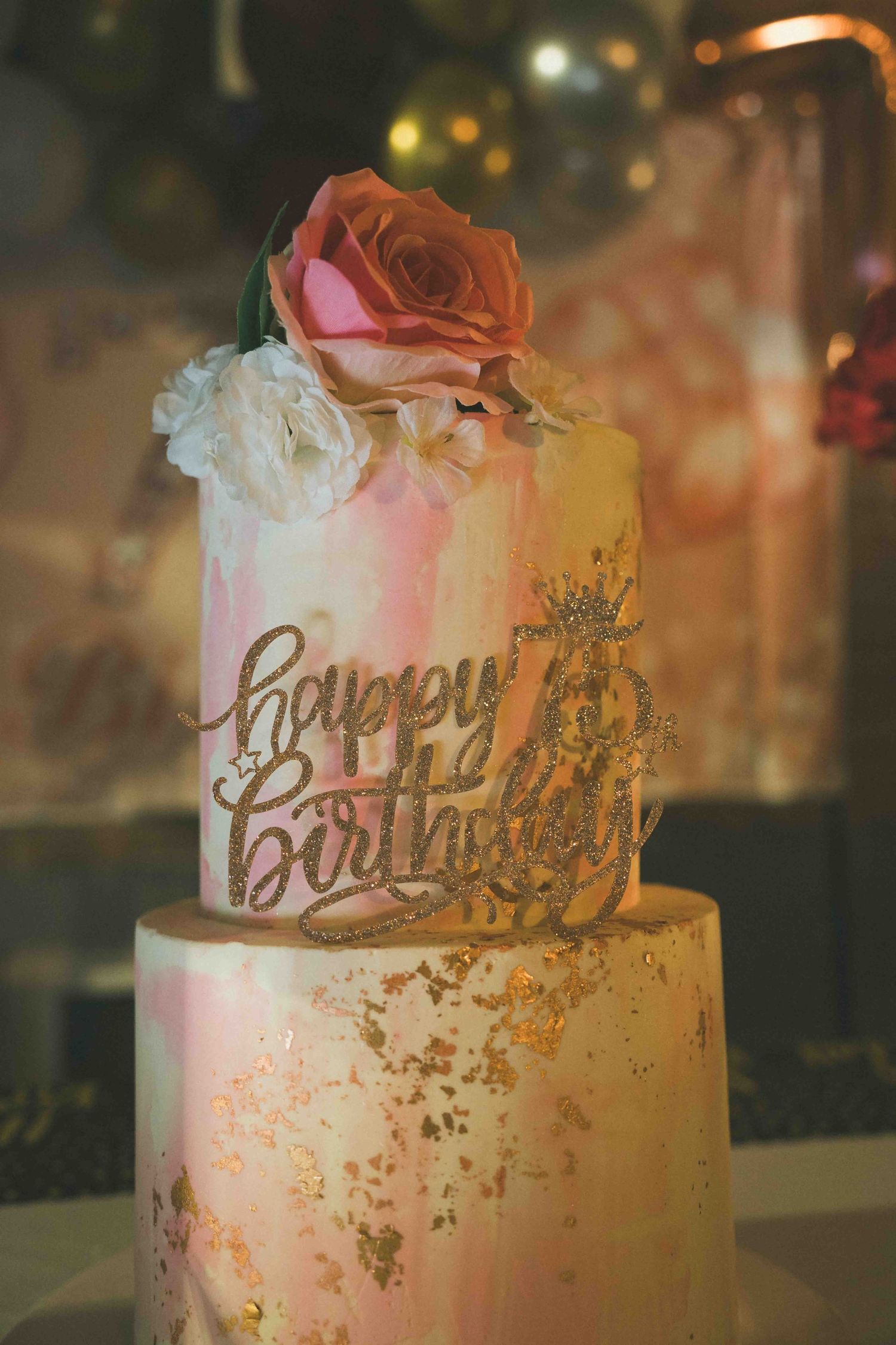 Wedding cake with gold accents, floral decorations and 'Happy Birthday' text in rose gold.