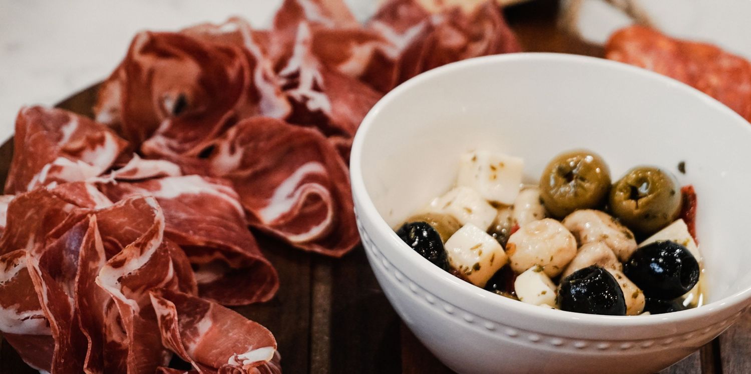 Thinly sliced prosciutto and cured meats arranged on a rustic wooden board with olives and cheese.